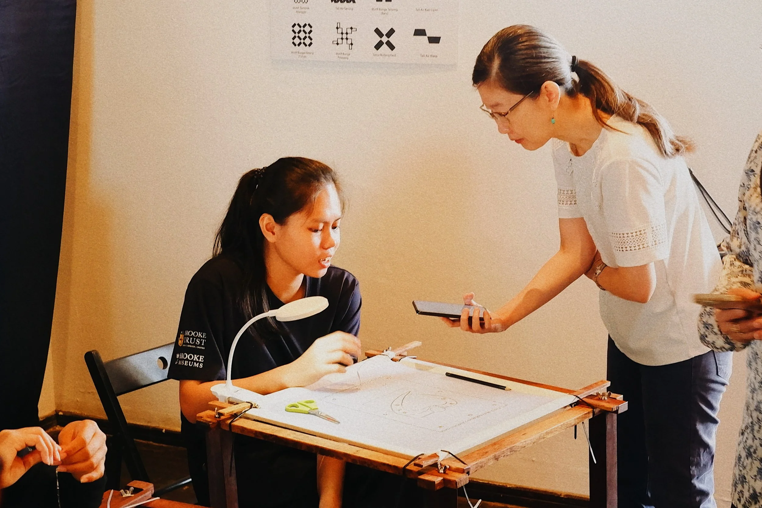 A woman in a white shirt is leaning over a table, showing something on her smartphone to a young woman seated at the table. The seated woman is wearing a black shirt and appears to be working on a craft or textile project, with a drawing, scissors, a