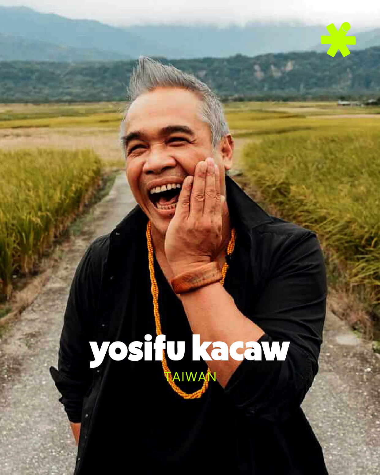 A man with gray hair laughing and touching his cheek with one hand, standing on a dirt path in a rural area with green fields and mountains in the background, wearing a black jacket and yellow beaded necklace. Text overlays: 'yosifu kacaw' and 'TAIWAN'.