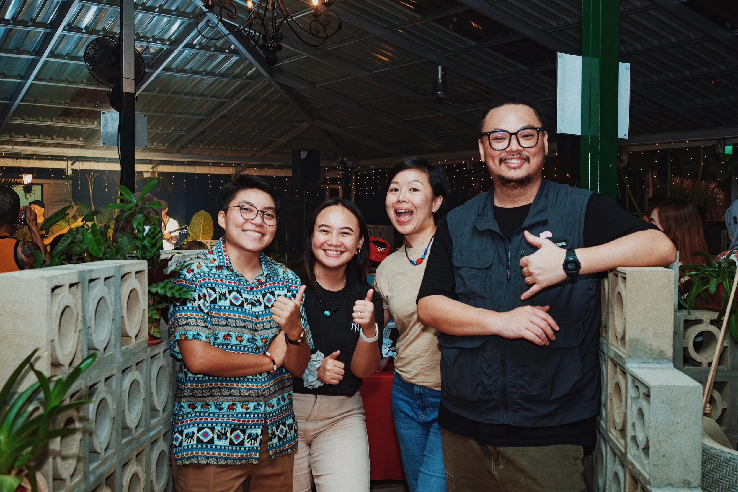 Four friends smiling and posing for a photo at a social gathering or party, standing indoors with decorative lights and plants in the background.