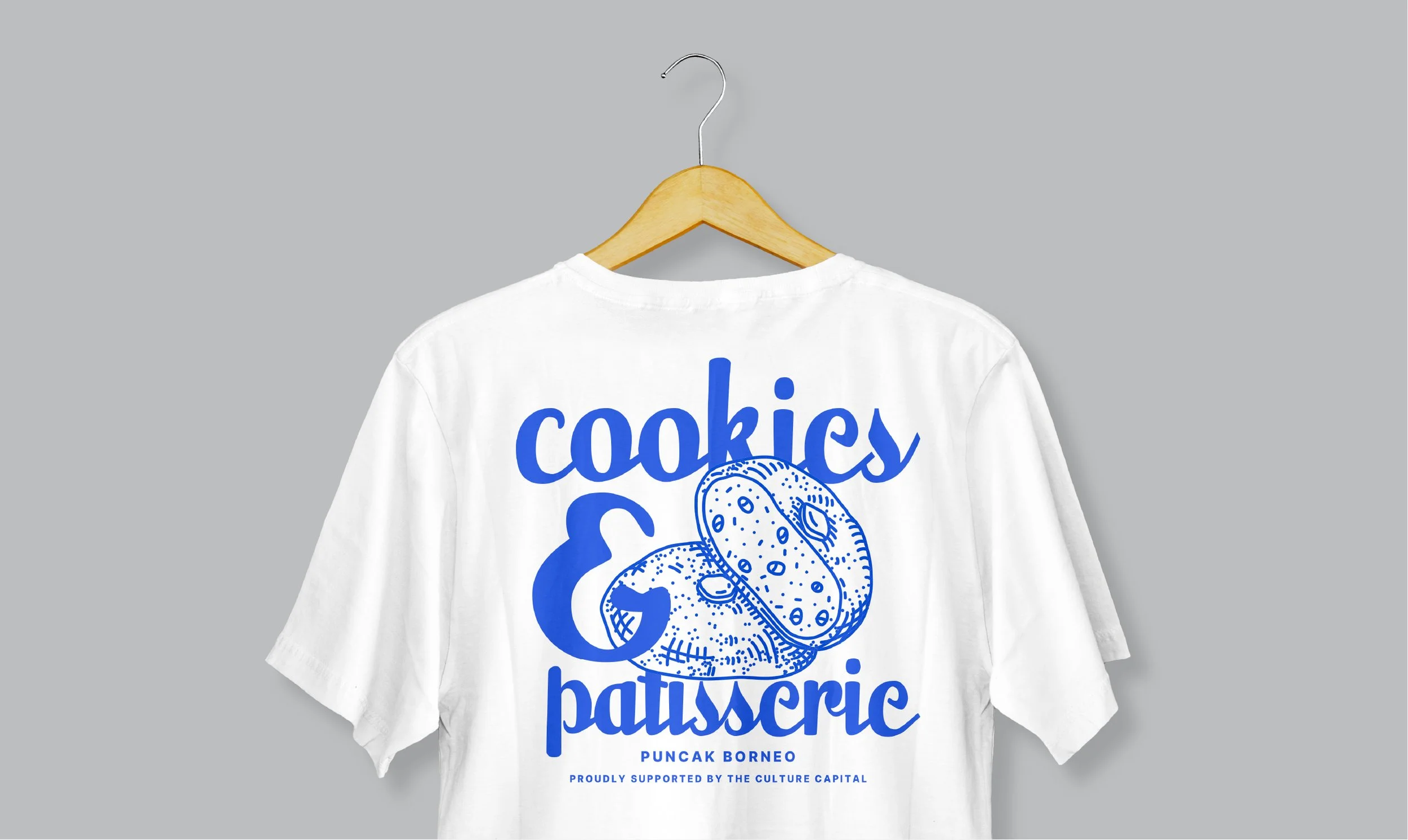 White T-shirt on a hanger with blue graphic text reading "cookies & patisserie" and an illustration of a cookie, with additional smaller text "Puncak Borneo" and "Proudly supported by the Culture Capital."