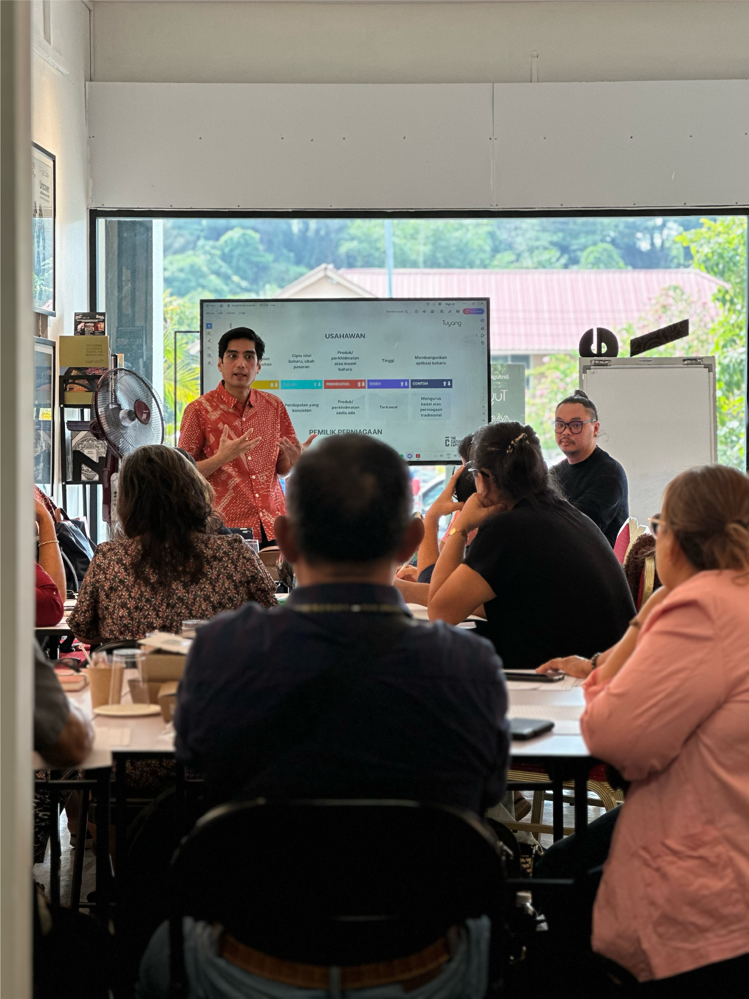 A man in a red shirt is giving a presentation to a group of people seated at tables indoors, with a large screen behind him displaying a slide with text and colorful sections.
