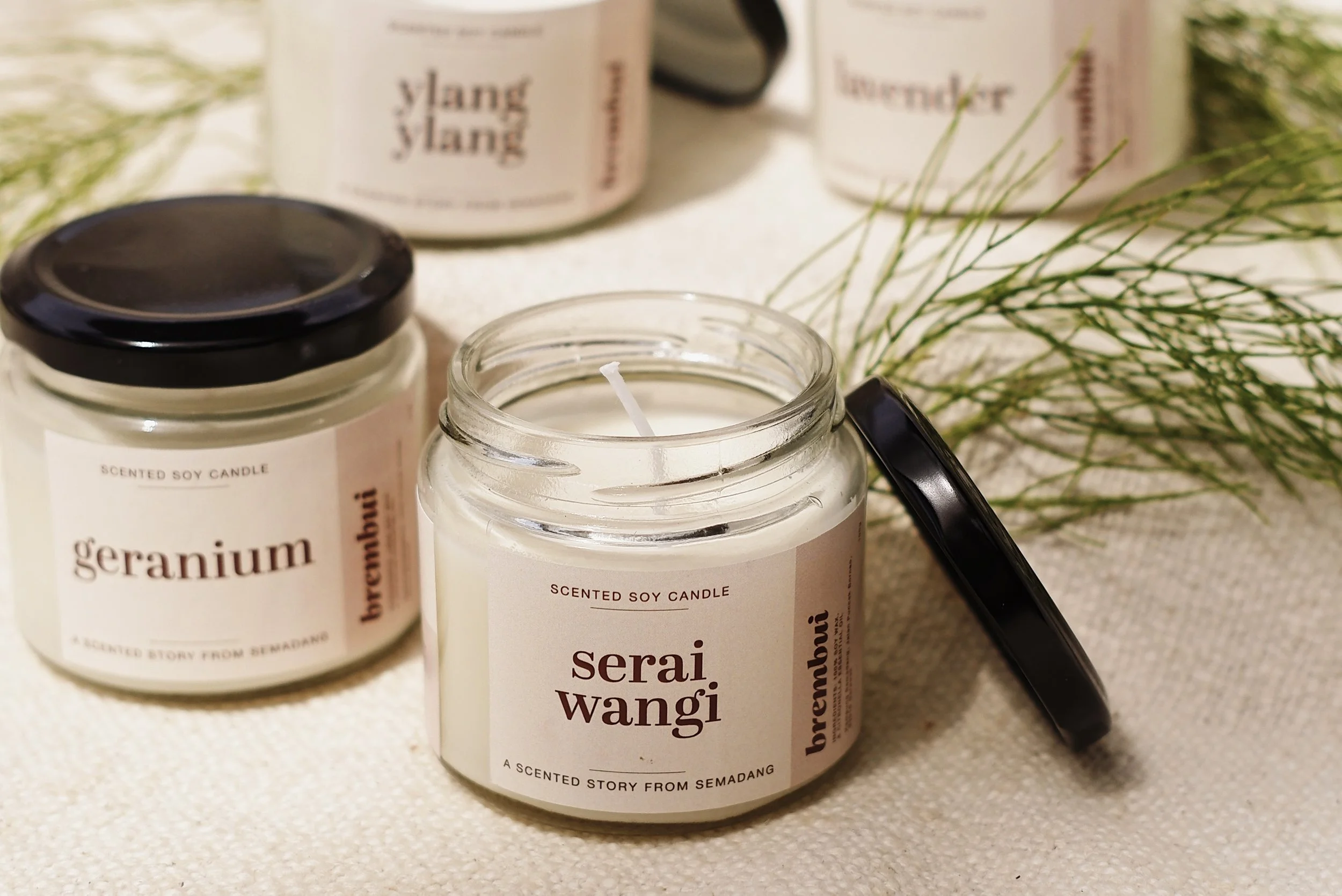 Scented soy candles in glass jars labeled 'serai wangi' and 'geranium', with some candles having black lids, placed on a light-colored fabric surface with greenery in the background.