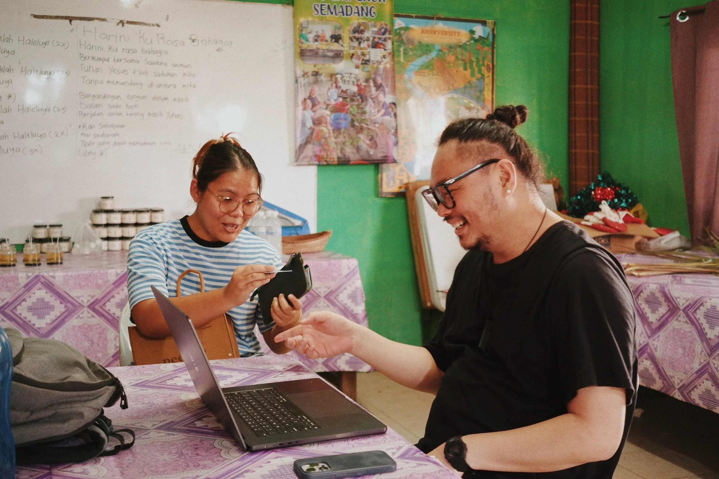 Two people sitting at a table with a laptop, sharing a moment of laughter, in a room decorated with posters and tablecloths with purple geometric patterns.