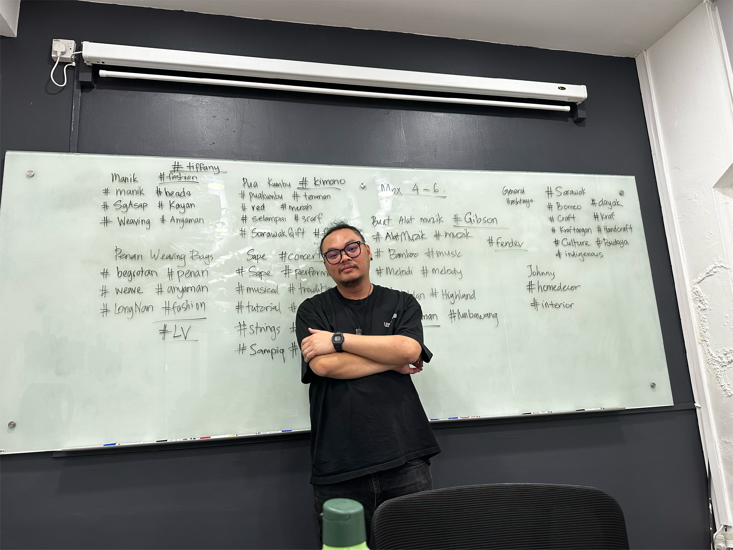 A man with glasses and a black shirt standing with arms crossed in front of a whiteboard filled with handwritten notes and hashtags, in a classroom or meeting room.