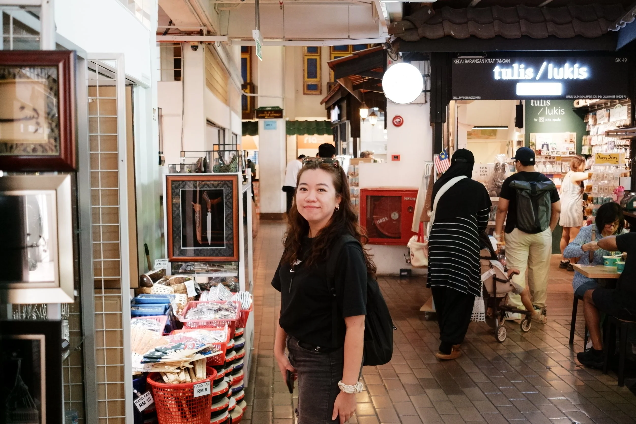 A woman standing inside a market or shopping area, smiling at the camera, surrounded by various merchandise and shoppers.