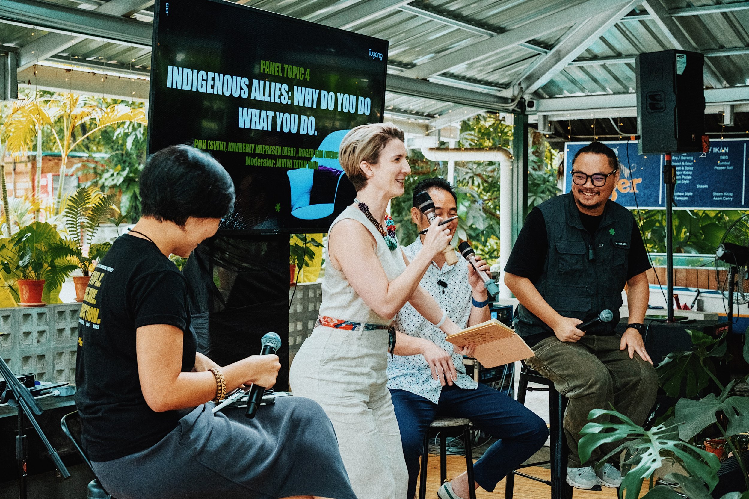 Four people seated and standing on a stage with potted plants in the background, participating in a panel discussion. A large screen behind them displays the topic 'Indigenous Allies: Why Do You Do What You Do.'
