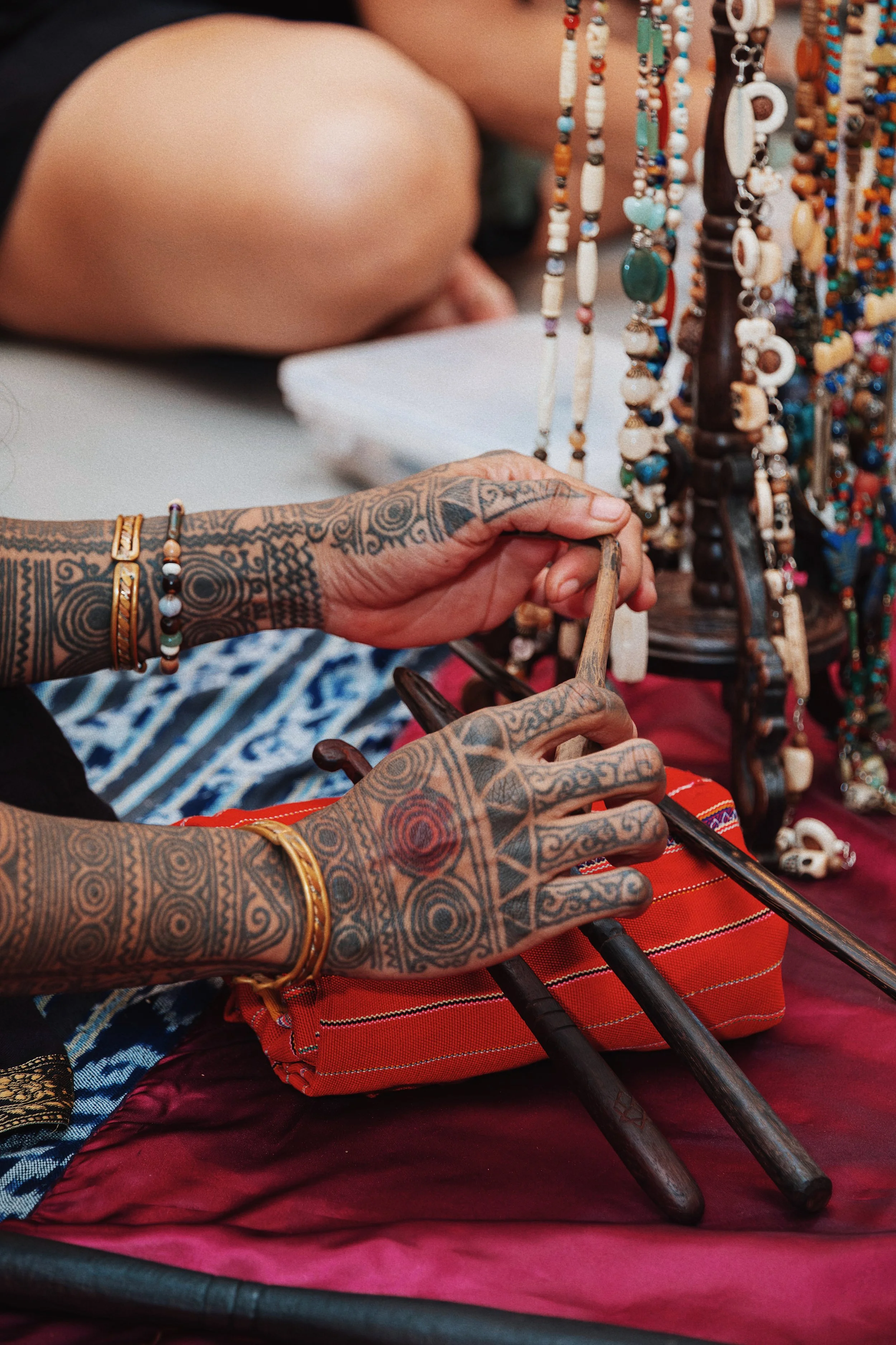 Close-up of heavily tattooed hands arranging traditional jewelry on display, with colorful beaded necklaces hanging nearby.