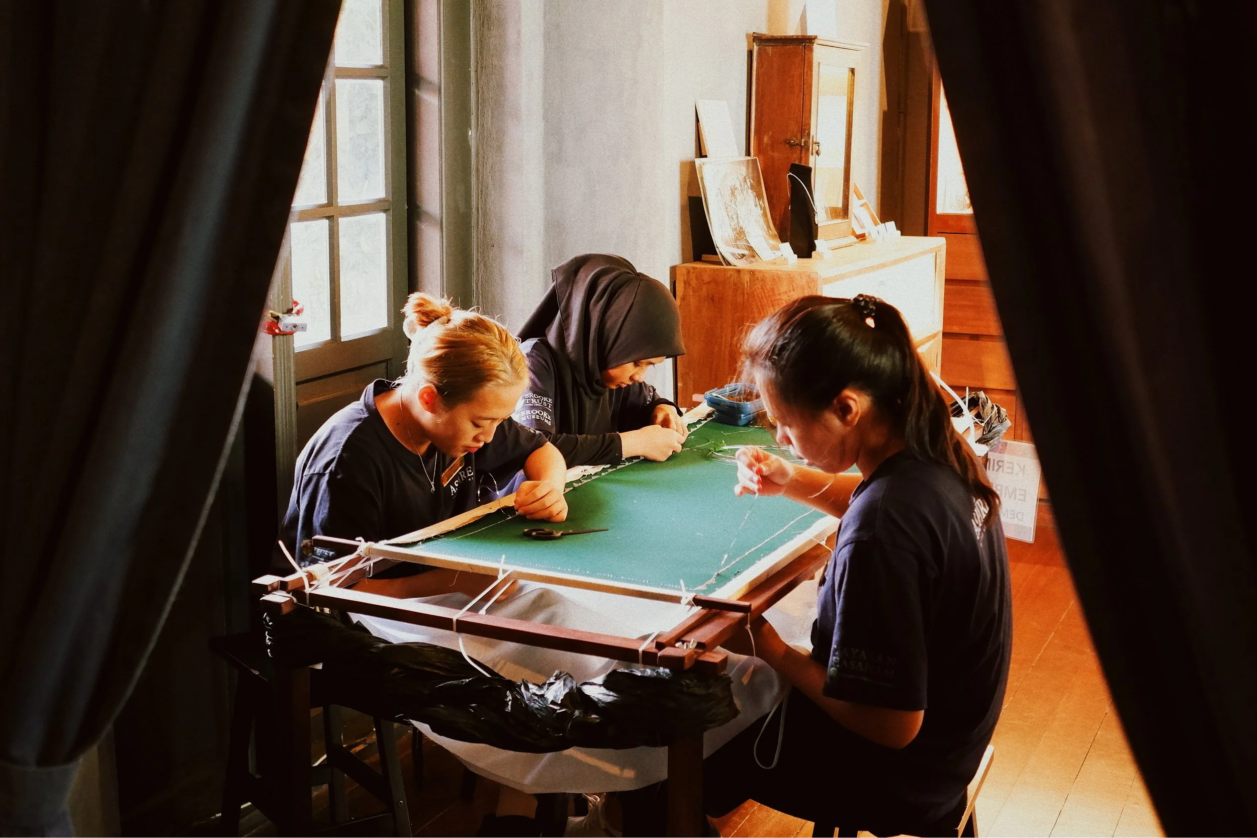 Three women working on a craft project at a table indoors, with craft materials and tools, seen through a partially opened curtain.