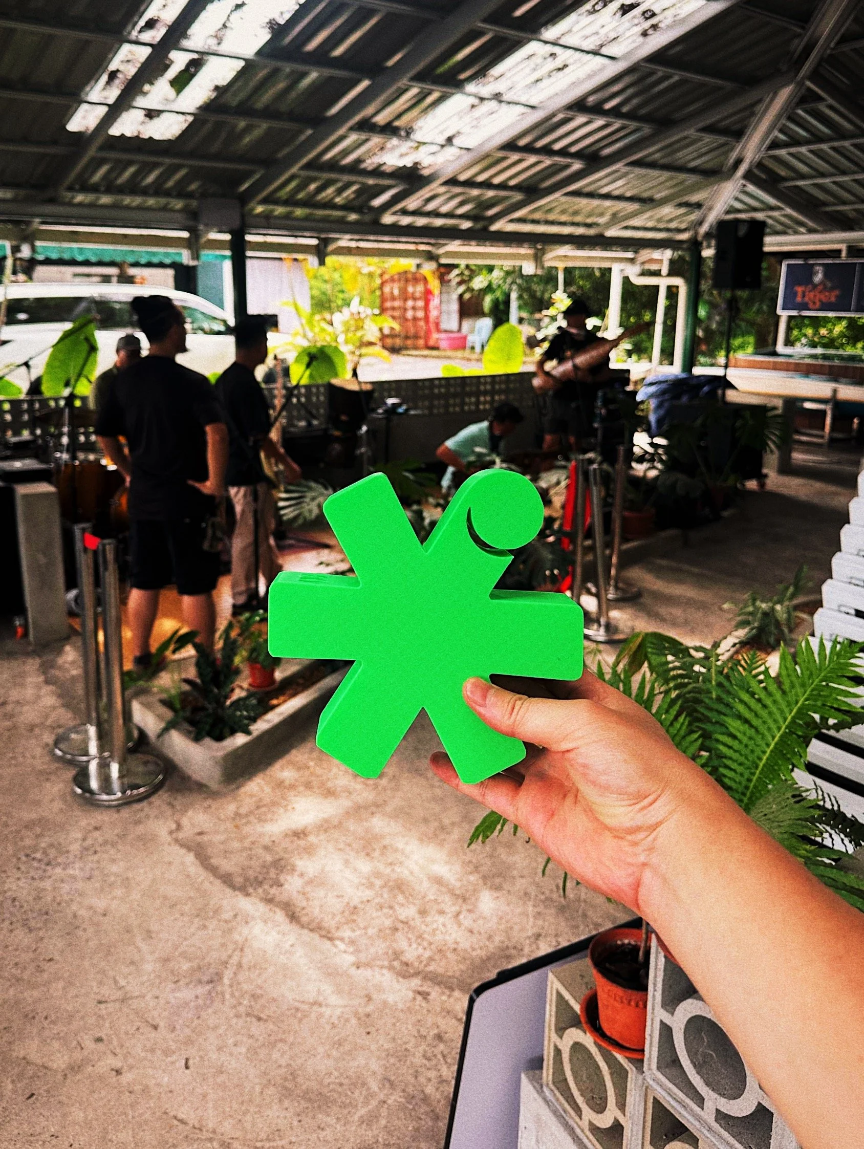 Person holding a bright green star-shaped sign with a small circle on top in an indoor market or dining area, with people in the background near plants and a metal roof.