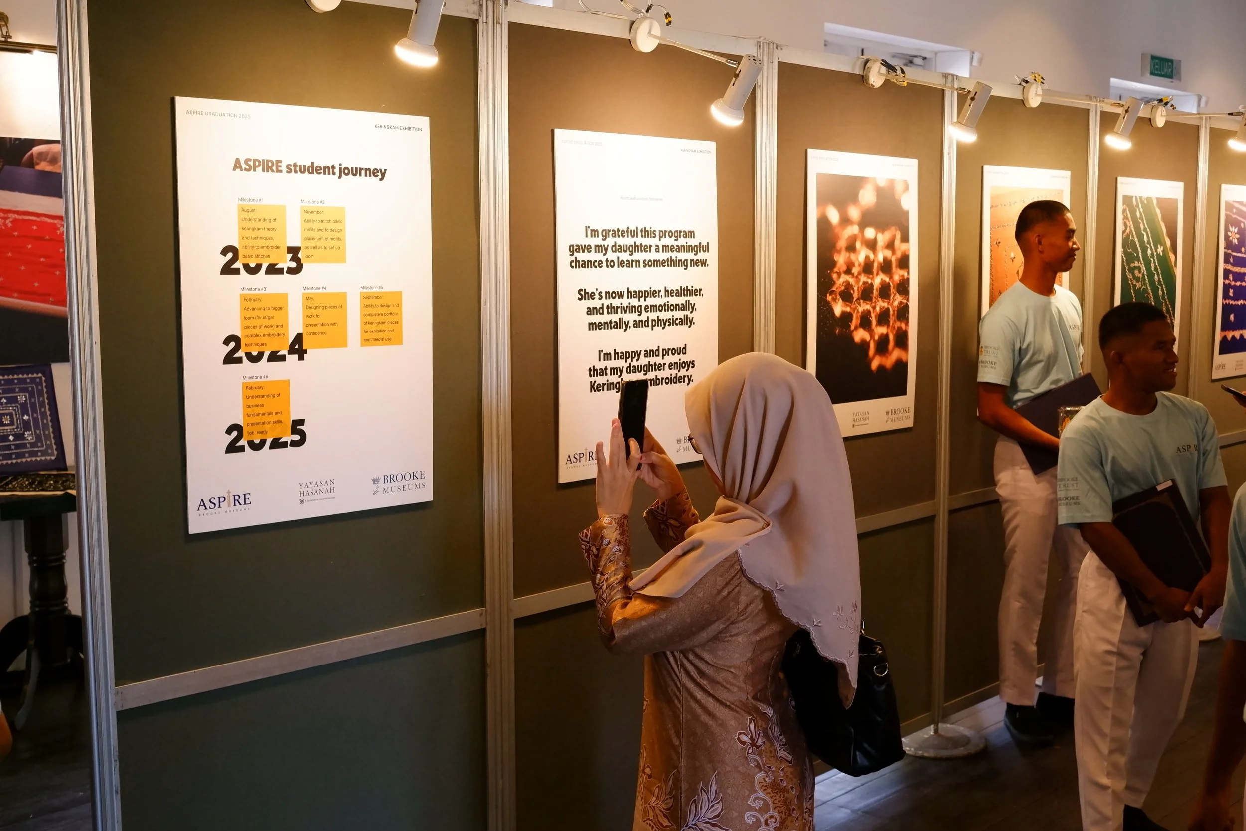 A woman wearing a beige hijab and brown patterned dress is taking a photo of an exhibition poster with her smartphone. She is surrounded by people in light blue shirts at an indoor art exhibition with framed posters and spotlights.