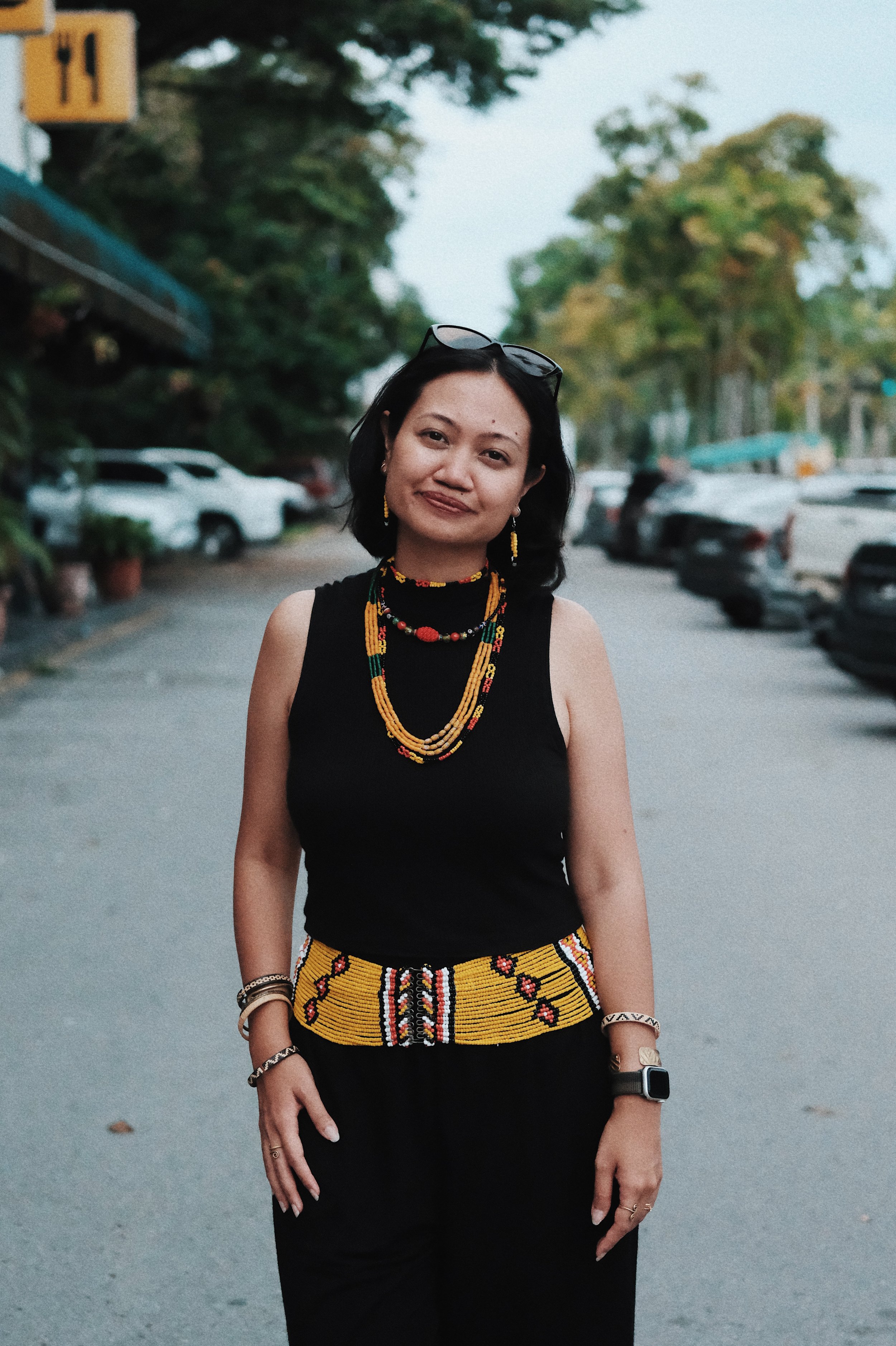 A woman standing outdoors on a parking lot road in the evening, wearing a black sleeveless top, black pants, colorful beaded jewelry, and sunglasses on her head, with trees and parked cars in the background.
