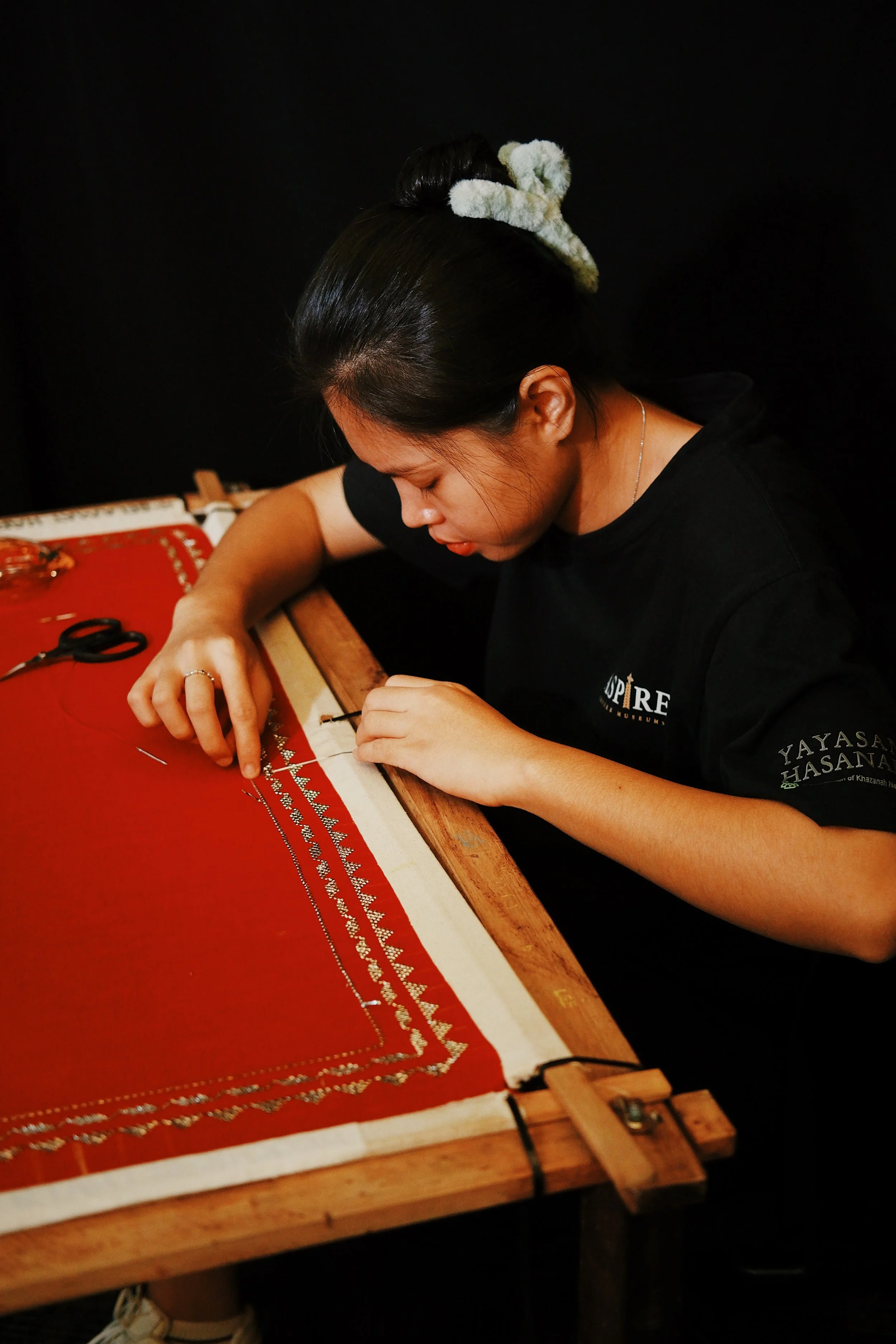 A woman working on intricate embroidery on a red textile, seated at a wooden frame with various tools nearby.