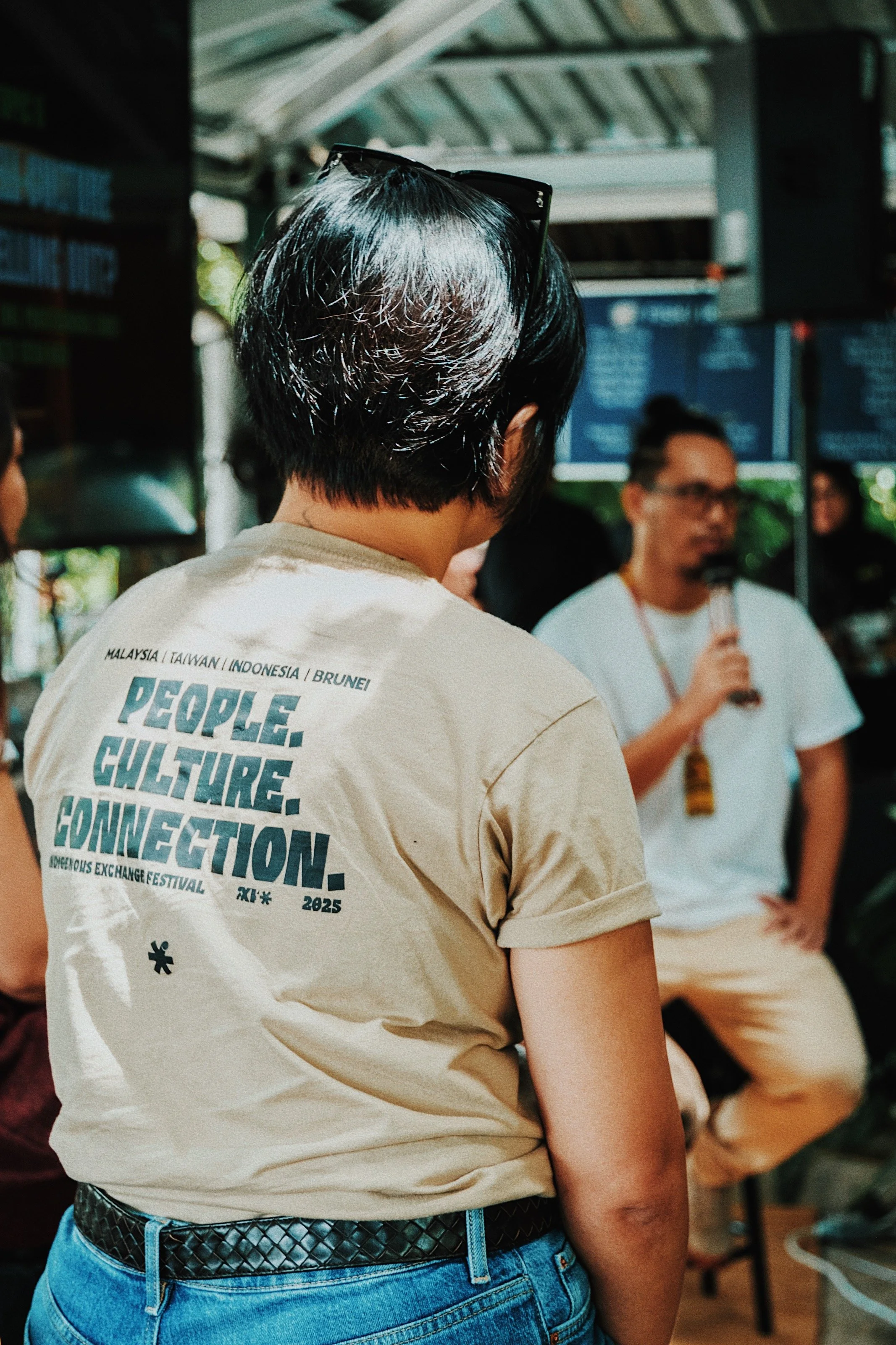 Person with black hair, wearing glasses on their head, a beige T-shirt with text about IndoPacific exchange festival, and blue jeans, facing a person speaking into a microphone at an outdoor event.