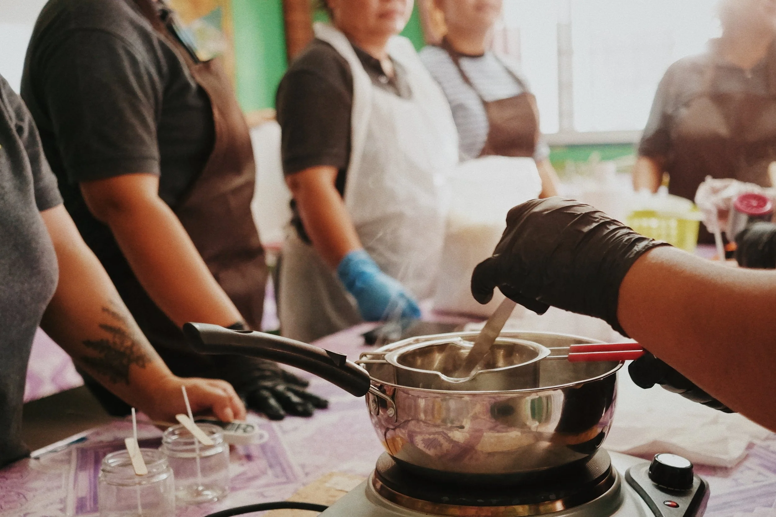 People in aprons and gloves participating in a cooking class, with a stove and a steaming pot in the foreground.