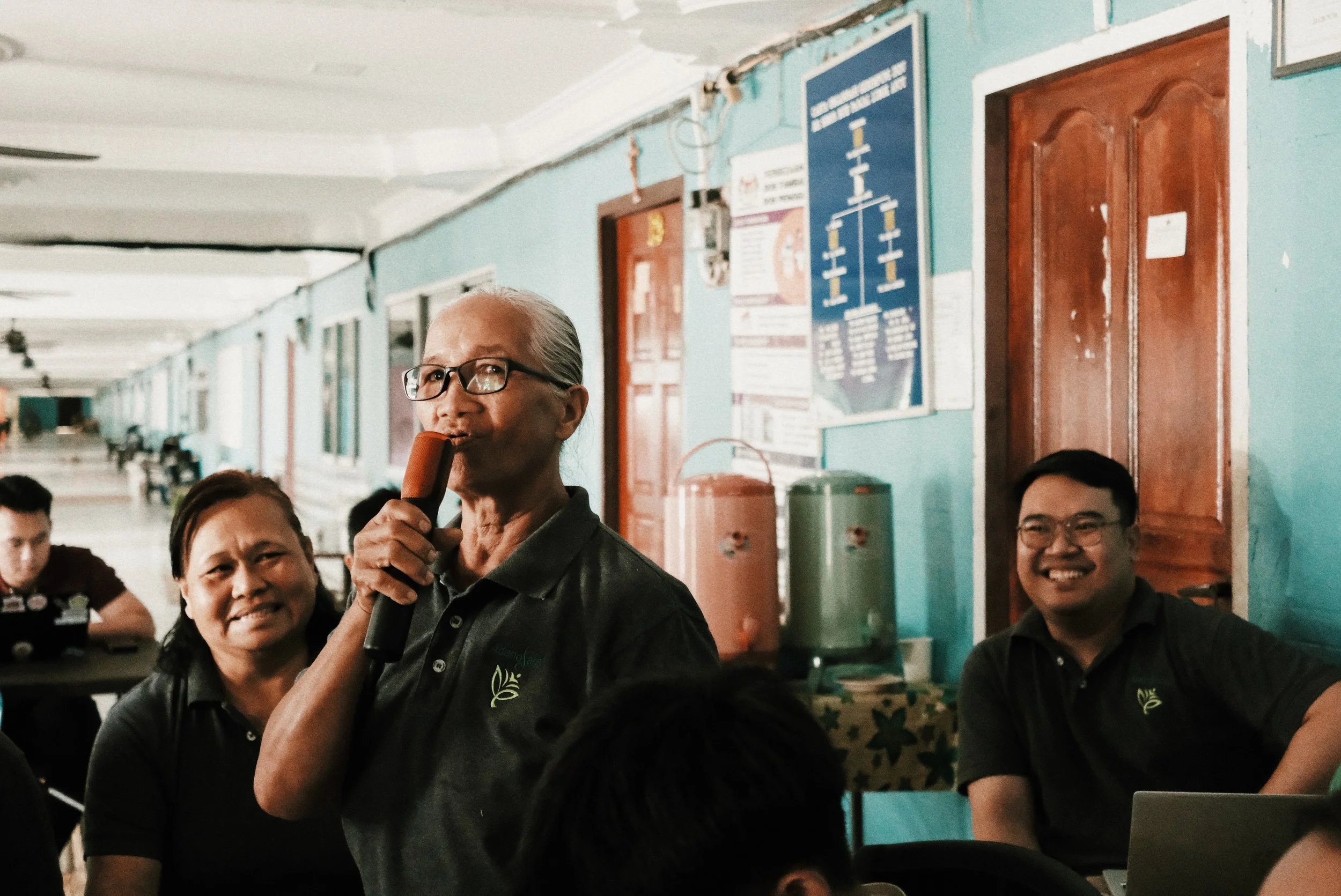 An older woman with gray hair and glasses holding a microphone, smiling, in a room with teal walls and other people seated around her.