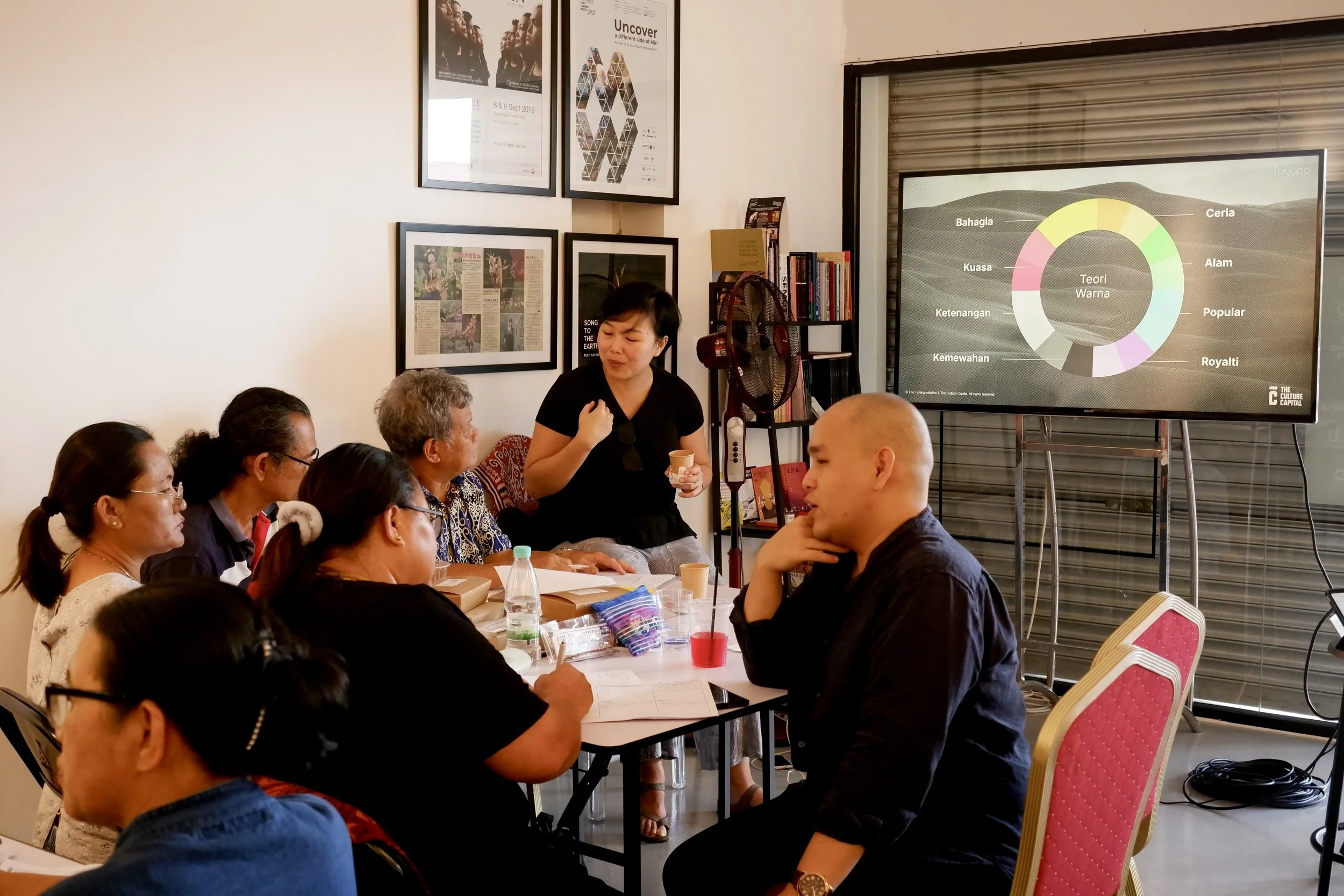 Group of people in a classroom or meeting room attending a presentation. A woman is standing and speaking to the group, with a display screen showing a colorful chart behind her. Several people are seated at tables, taking notes or listening attentiv
