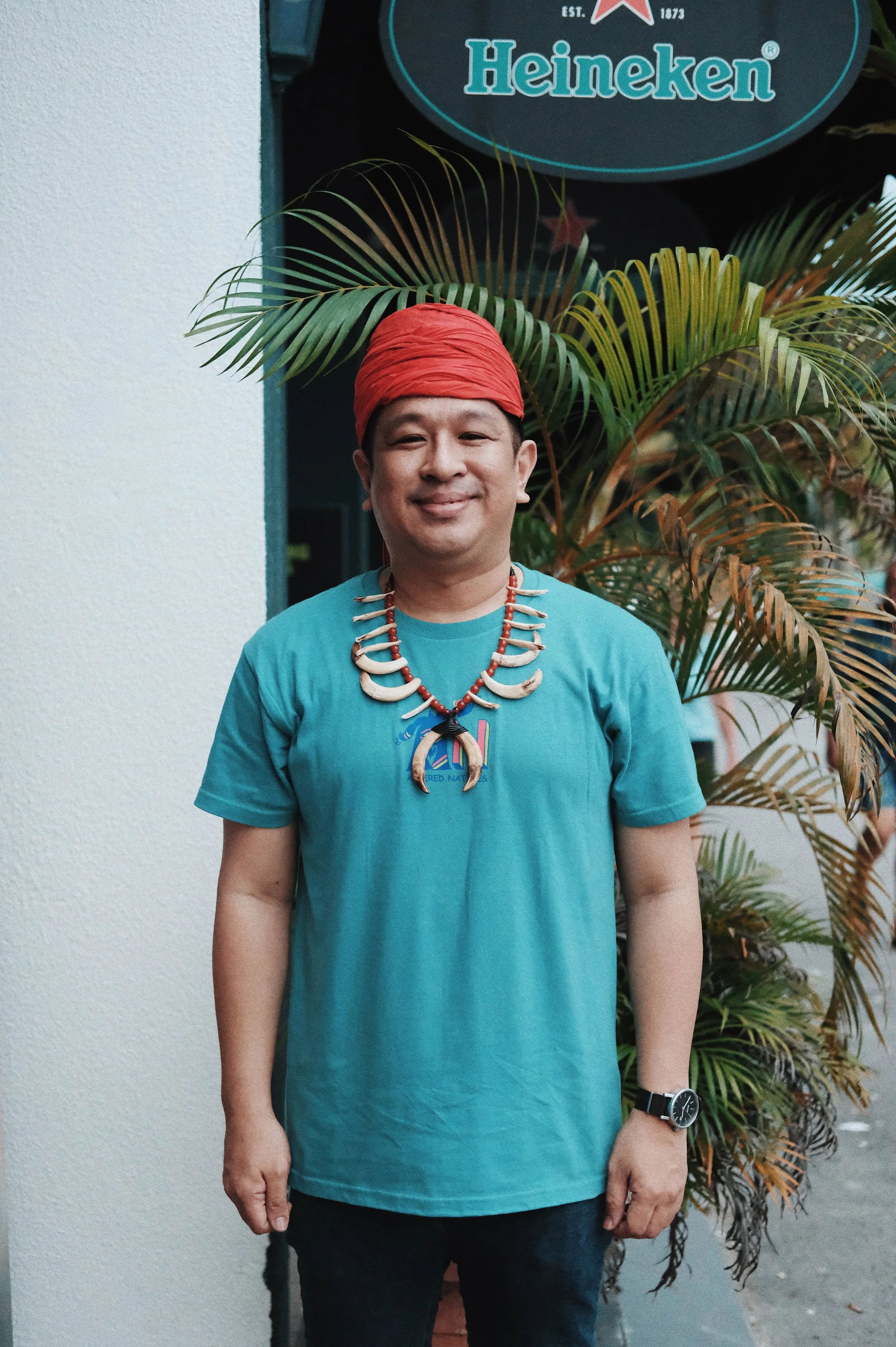 Man wearing a red headwrap and a beaded necklace with animal teeth, standing outdoors near a white wall and green plants, with a Heineken sign in the background.