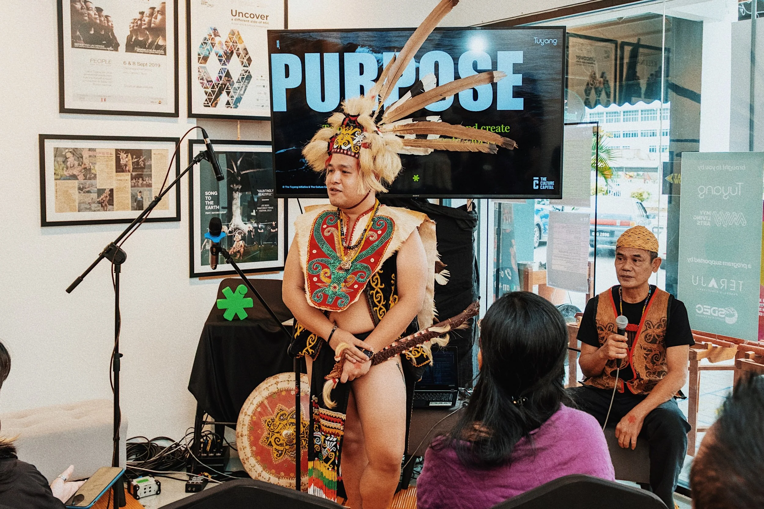 A man wearing traditional indigenous attire, including a headdress with feathers and a decorative vest, is speaking into a microphone at a cultural event. A woman with a woven hat is sitting nearby, and there are audience members listening in a room 