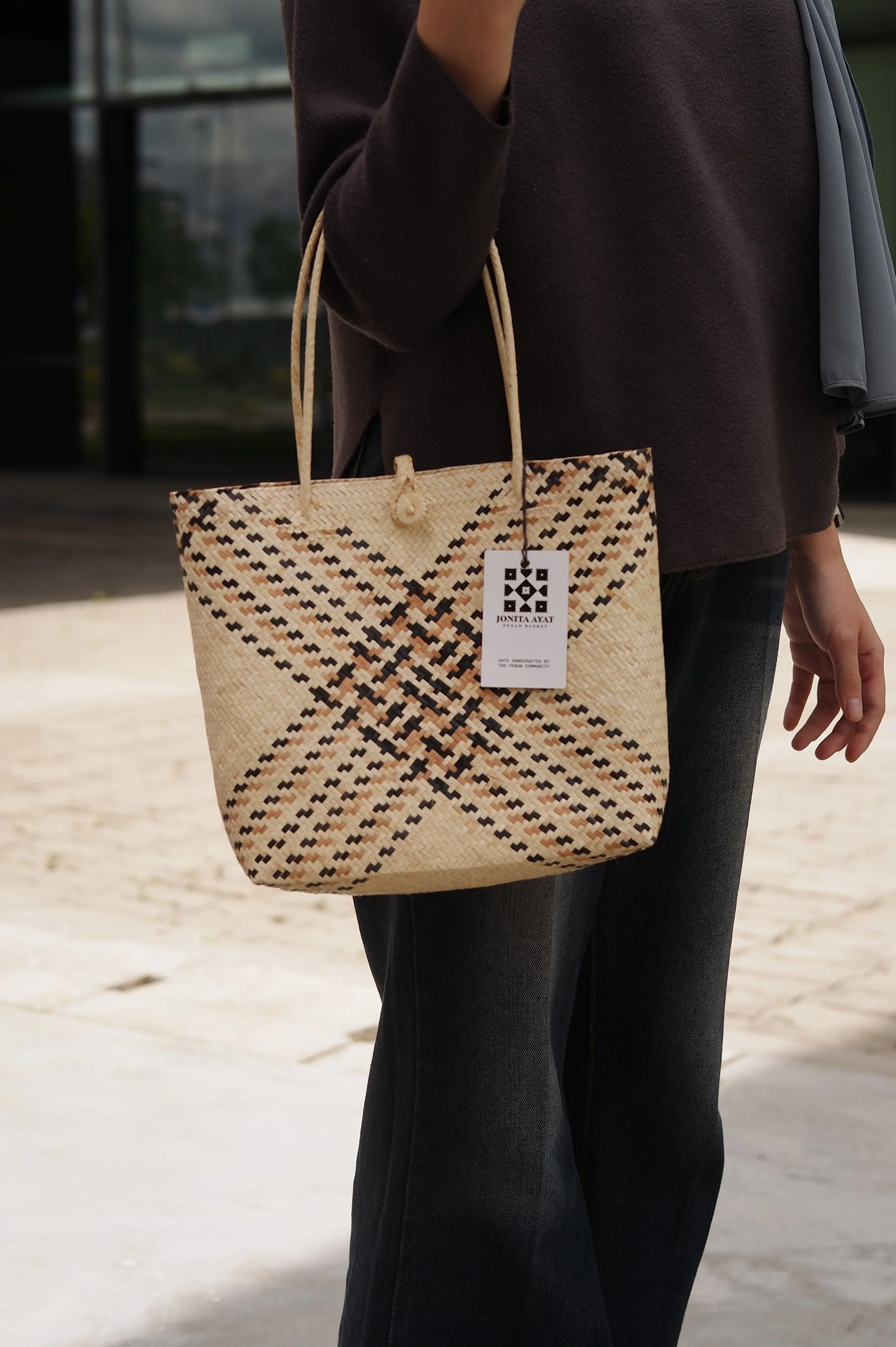 A person holding a woven beige tote bag with black and brown geometric patterns, wearing a dark brown sweater and black jeans, standing outdoors.