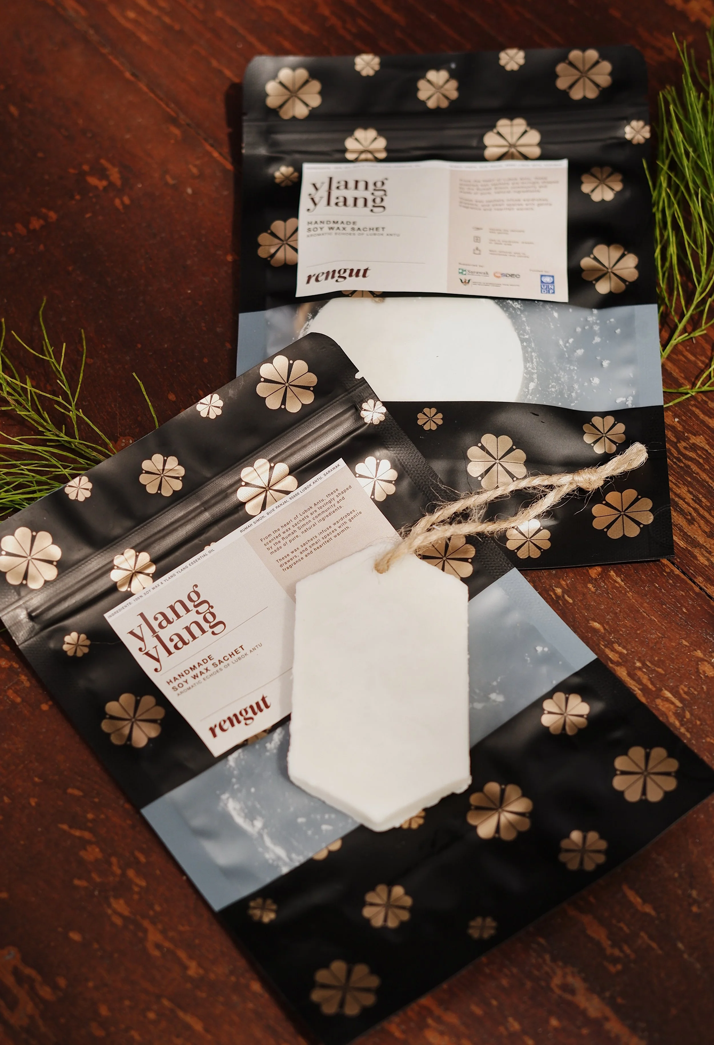Two black packaging bags with beige floral patterns and labels that read 'Ylang Ylang', 'rengut', and 'Handmade Soy Wax Sachet'. A white soy wax sachet with a string tied through a hole is placed on top. Green foliage is visible on the sides, and the items are on a wooden surface.