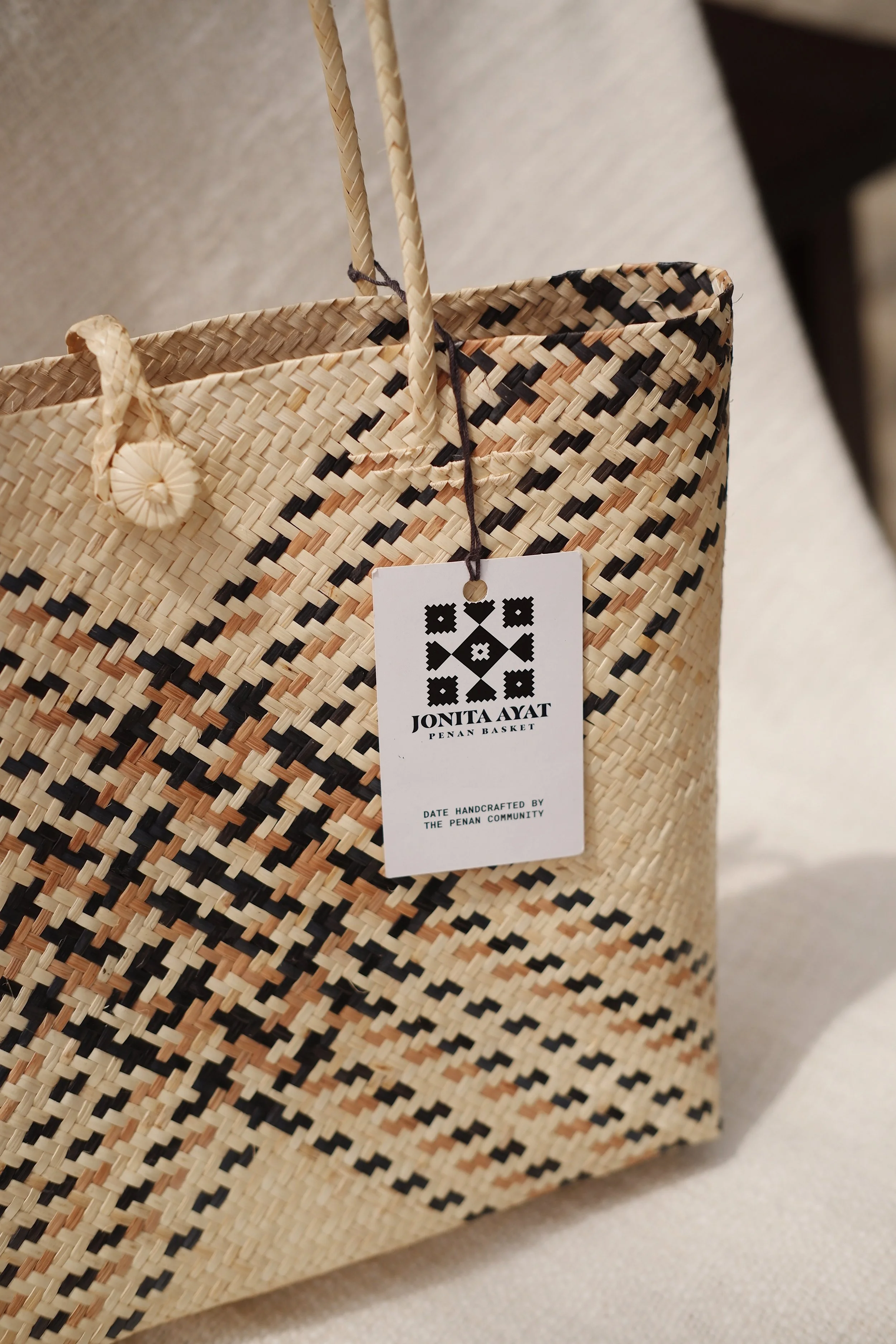 A woven straw basket with black and natural colored patterns, featuring a hanging tag that reads "Jonita Aayat Penan Basket" and "Date handcrafted by the Penan community."