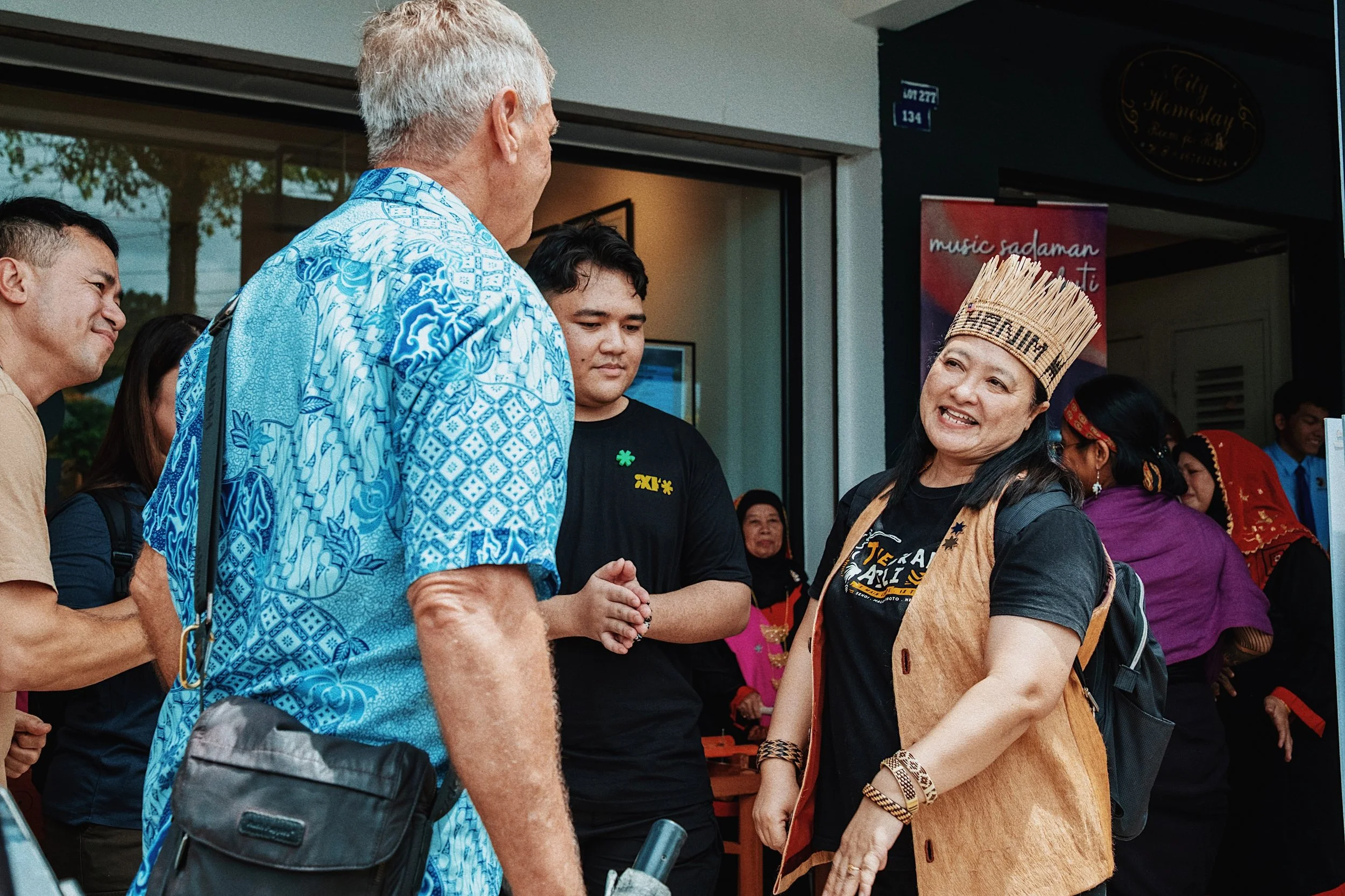 A woman wearing a traditional headdress and a vest, smiling and talking to a group of people, including a man in a blue patterned shirt, outside a building with people in the background.