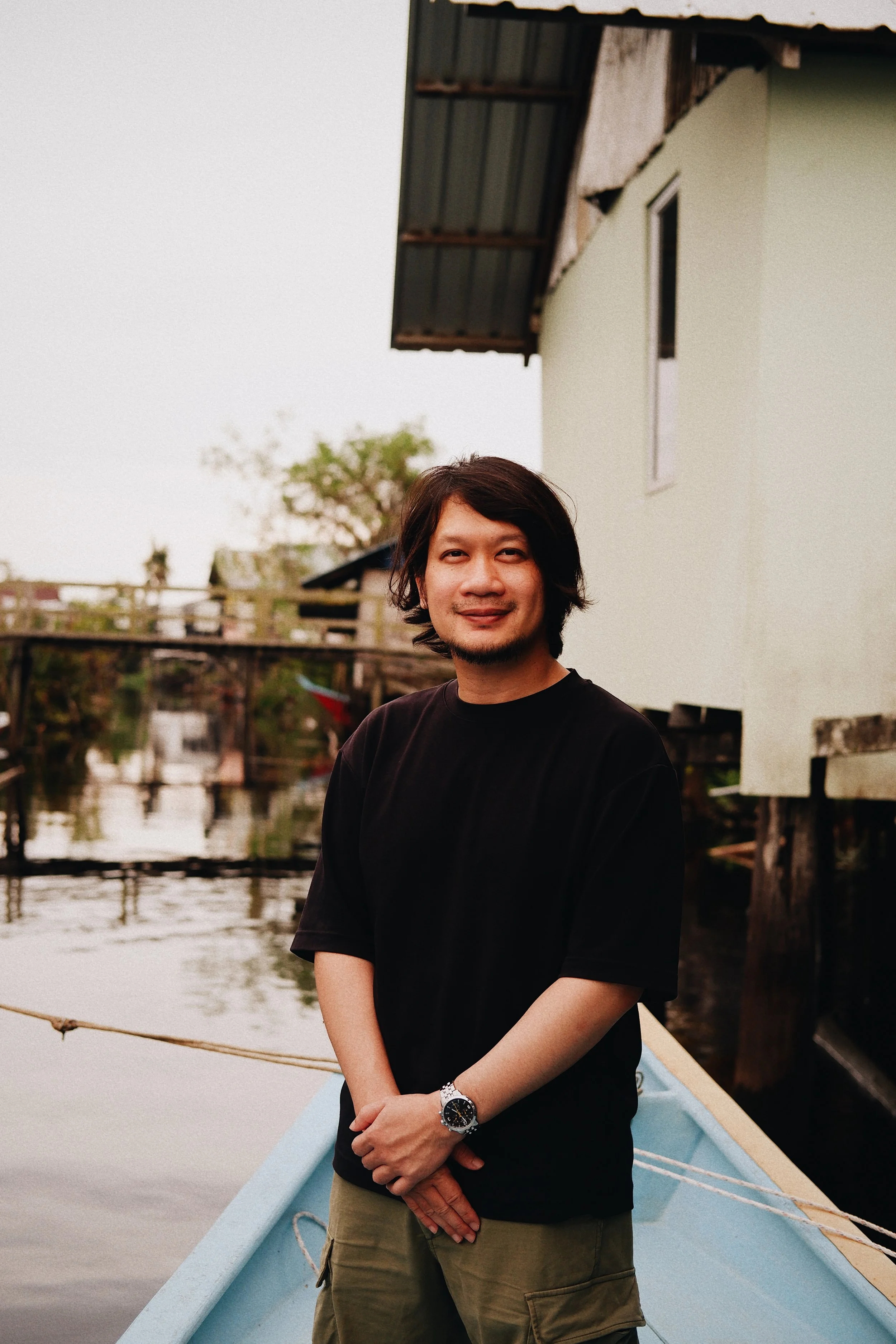 A man with shoulder-length black hair and a beard, wearing a black T-shirt and khaki shorts, standing on a boat near water with houses on stilts in the background.