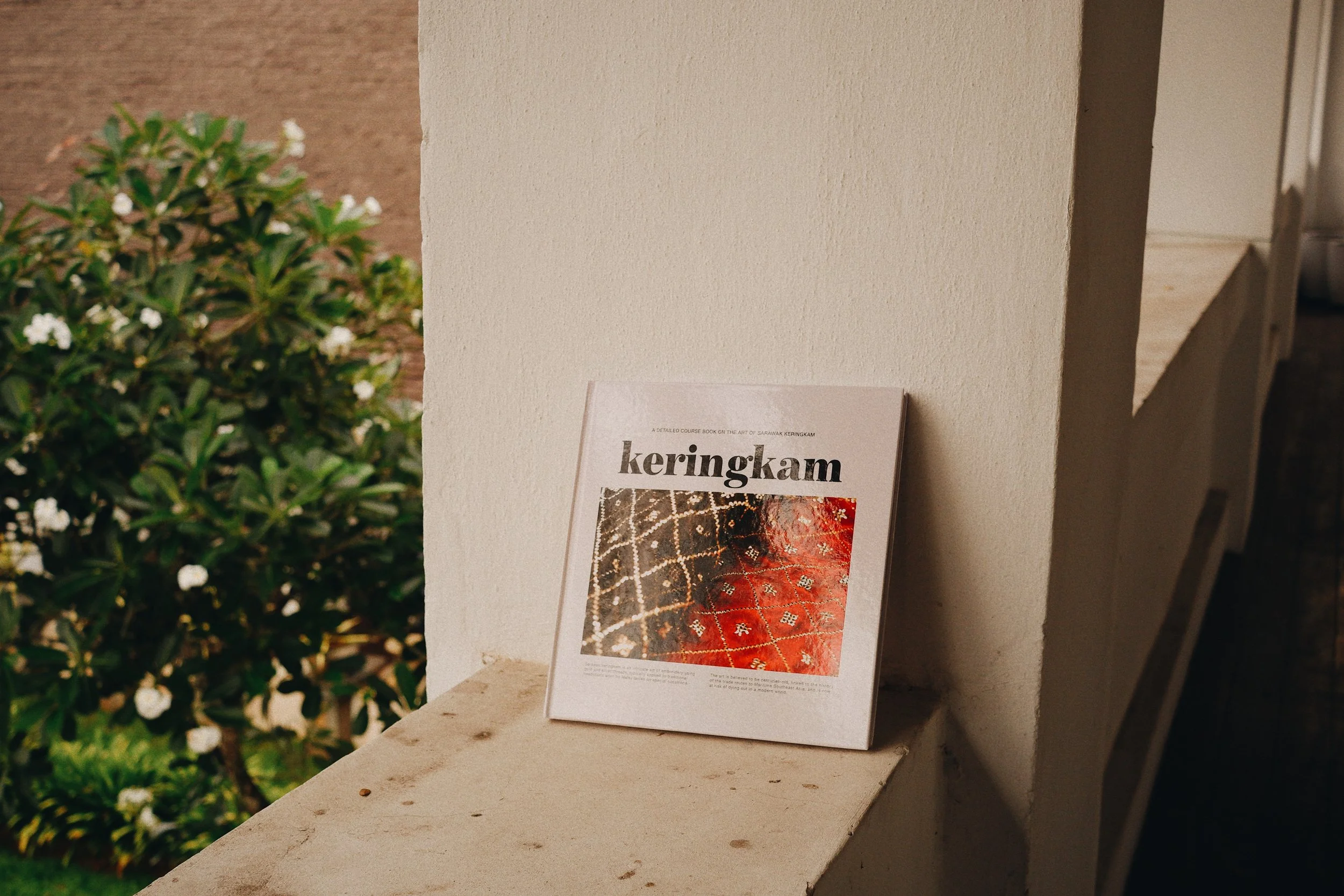 Book titled 'Keringkam' leaning against a beige wall on a ledge, with a plant with white flowers in the background.