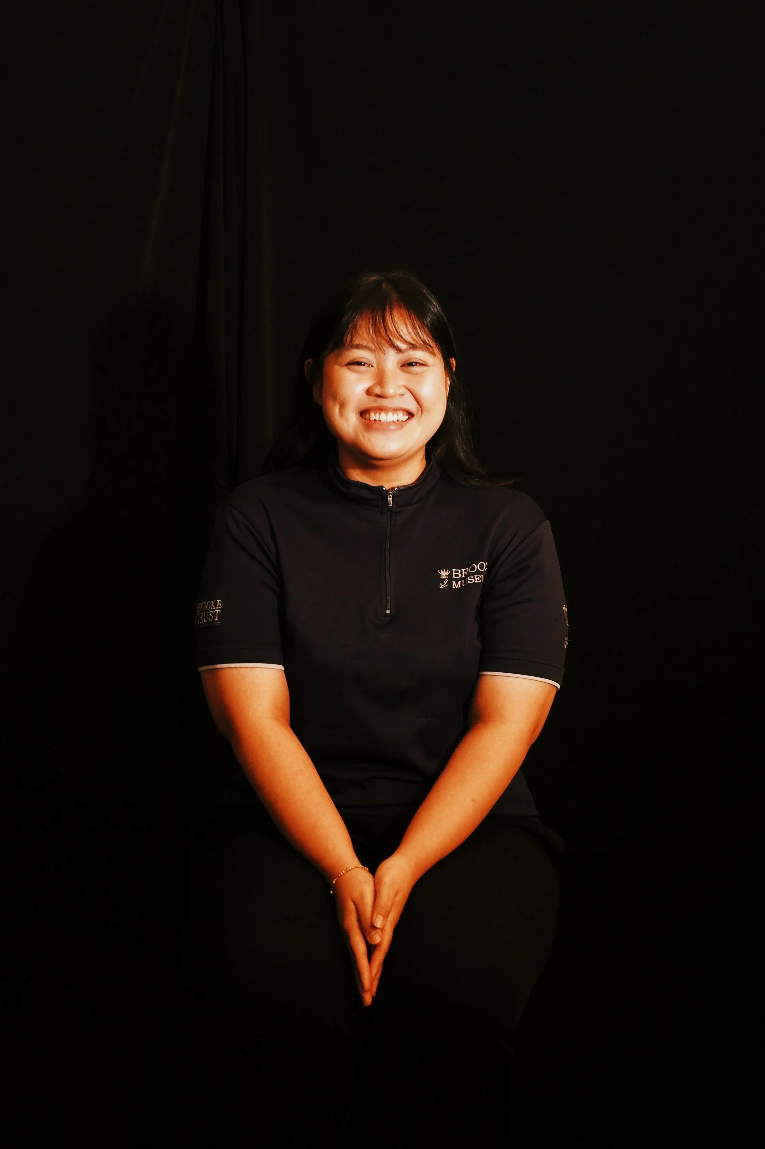 A smiling woman sitting against a black background, wearing a black polo shirt with a partially visible logo.