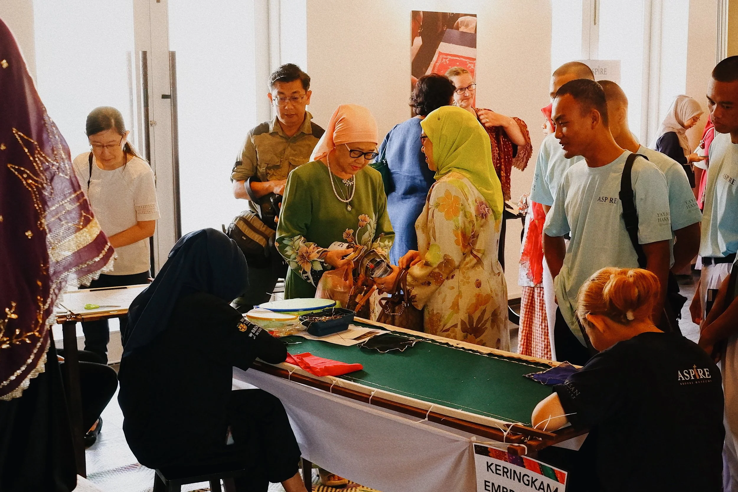 People gathered around a table at an indoor cultural event, with some examining fabric and accessories, while others stand nearby watching and chatting. The setting suggests a traditional or craft fair.