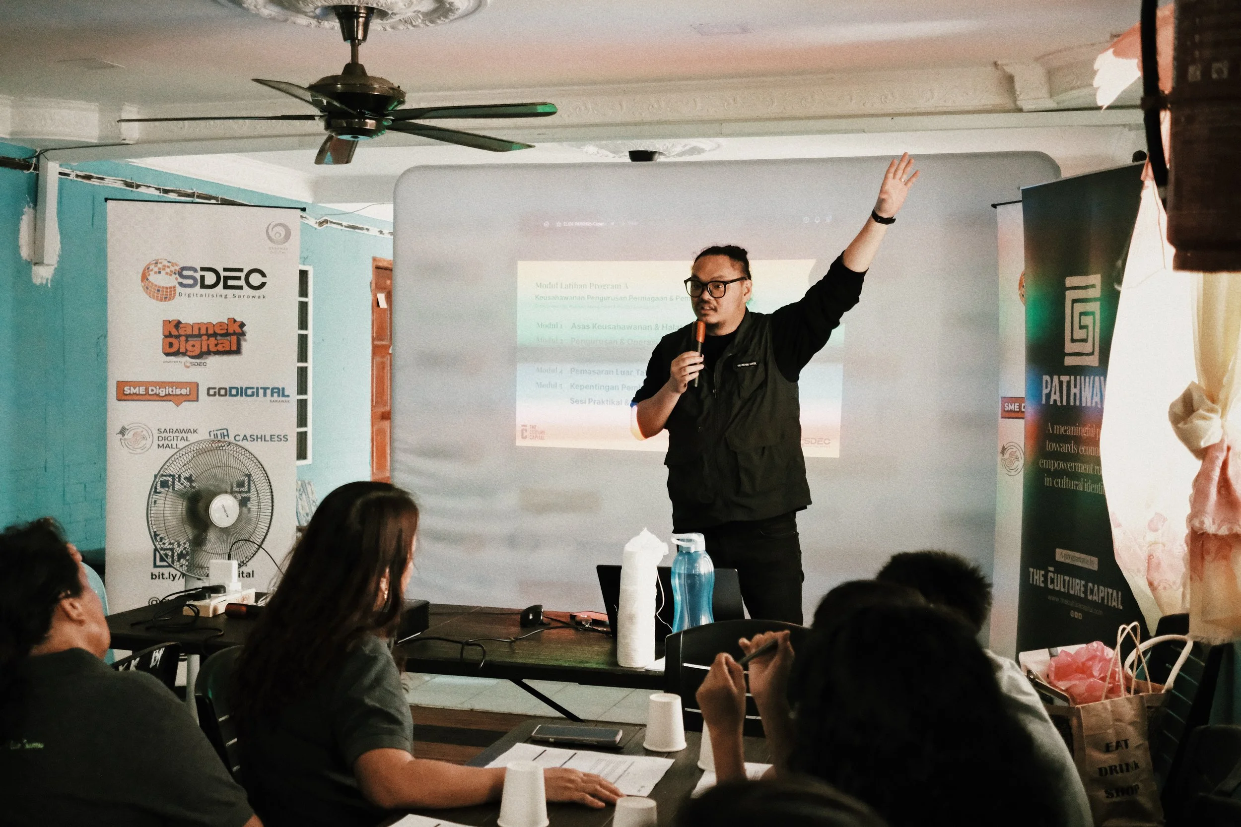 A man with glasses, wearing a black vest, is giving a presentation in a room with a white screen behind him. He is holding a microphone and raising his right hand. Several people are seated at a table, listening and taking notes. There are banners on each side of the presentation area.