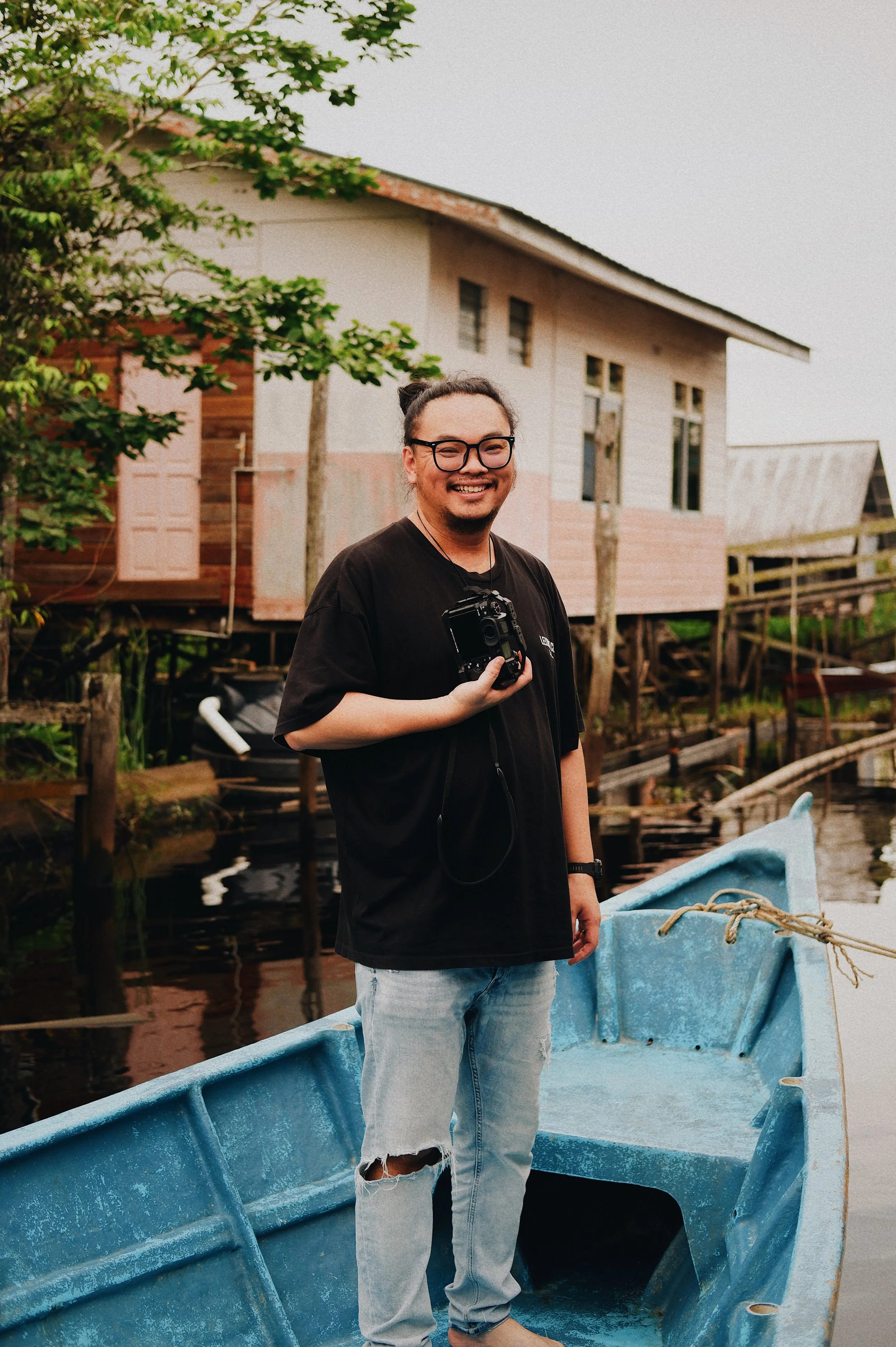 A smiling man with glasses, wearing a black t-shirt and ripped jeans, standing in a small blue boat near water, holding a camera, with a wooden house on stilts and greenery in the background.