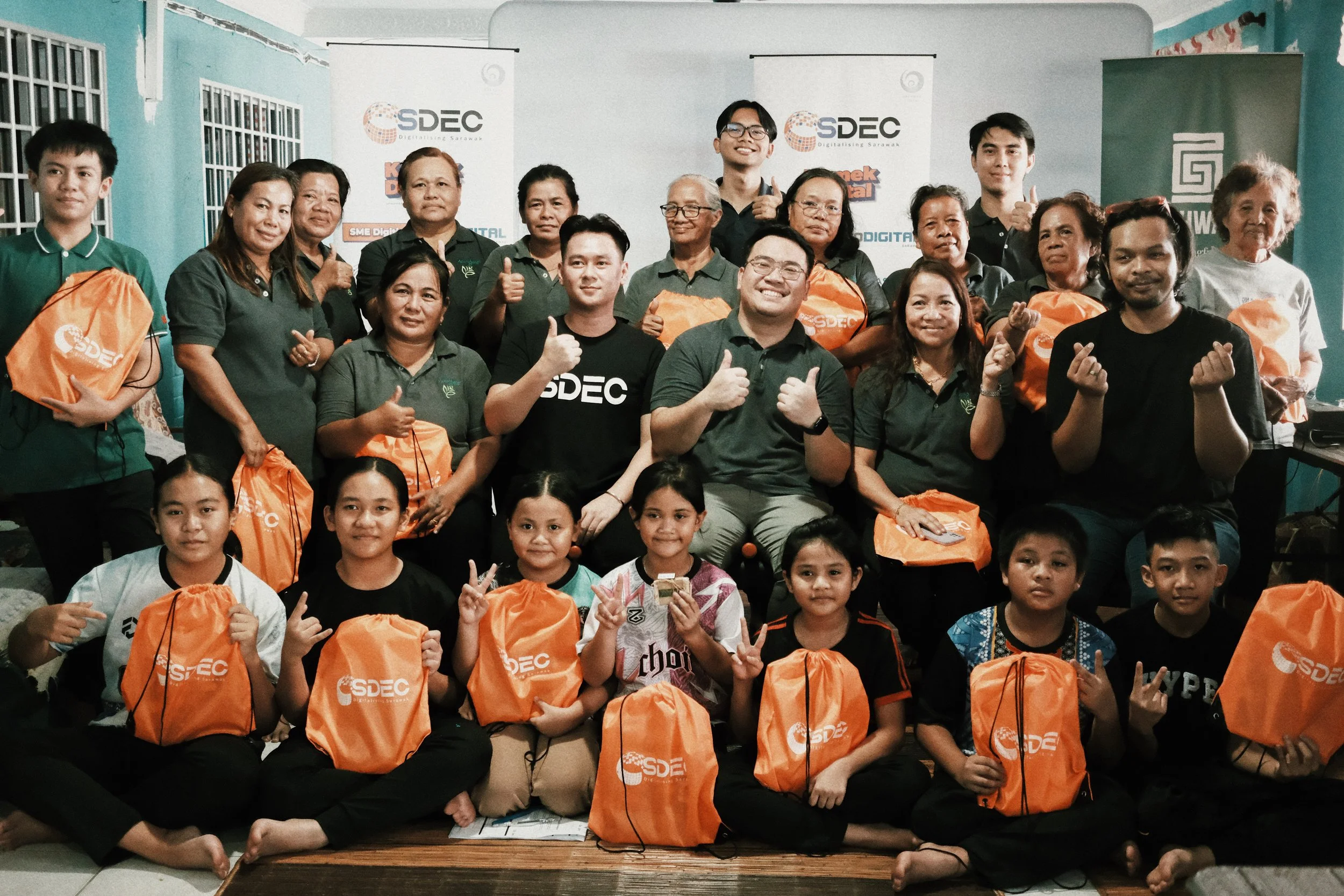 Group of children and adults posing indoors, holding orange bags with 'SDEC' logo, some making hand gestures, in front of banners with 'SDEC' branding.