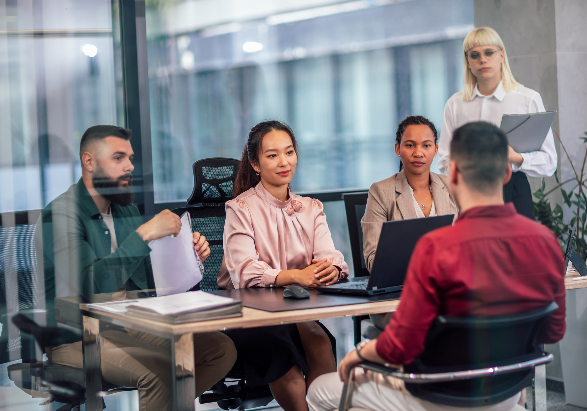 A diverse group of five business professionals in a meeting room with glass walls, one woman standing holding a folder, four people seated at a table with laptops and documents.