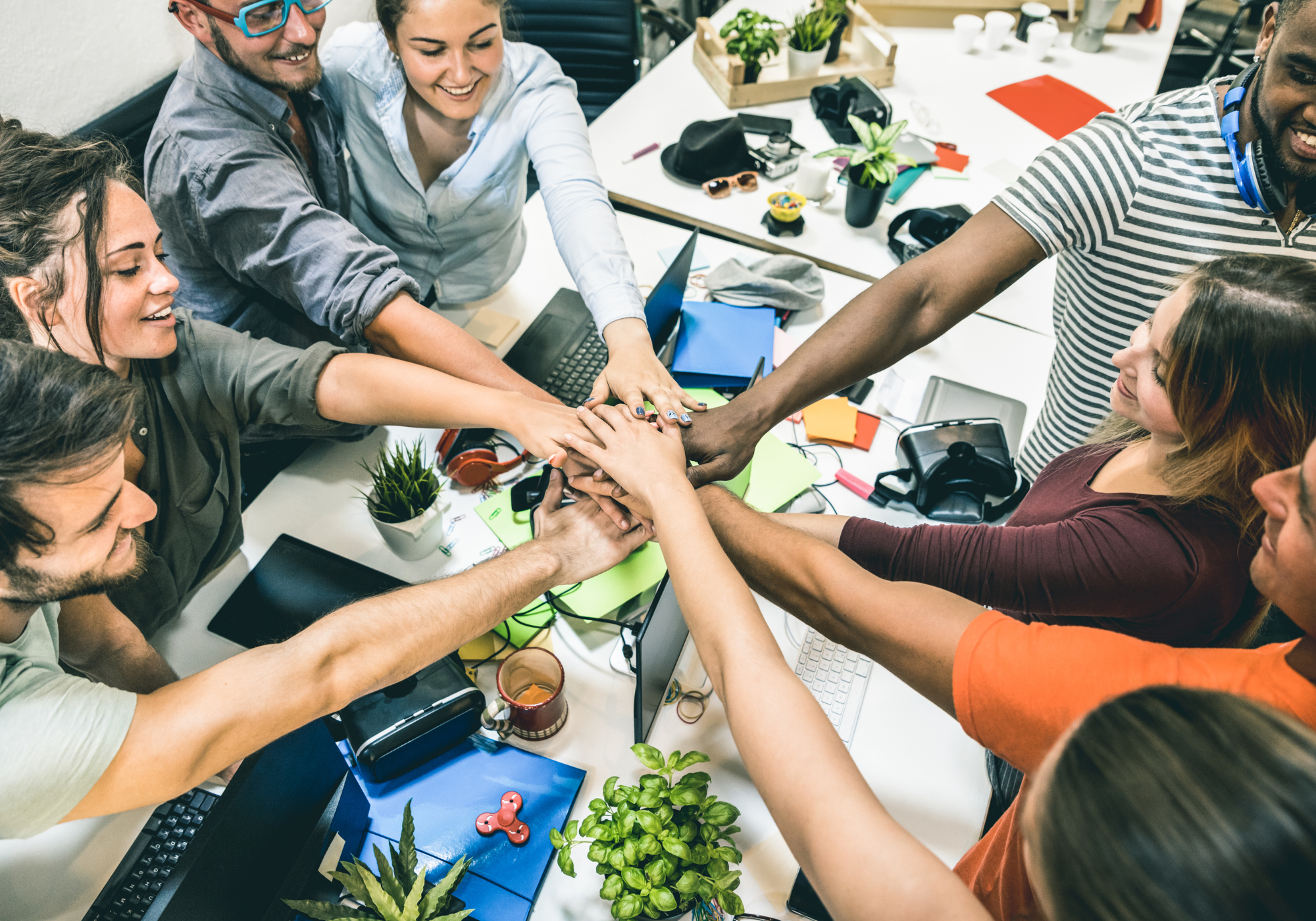 A diverse group of people in a workspace stacking their hands together in a team gesture surrounded by computers, plants, and office supplies.