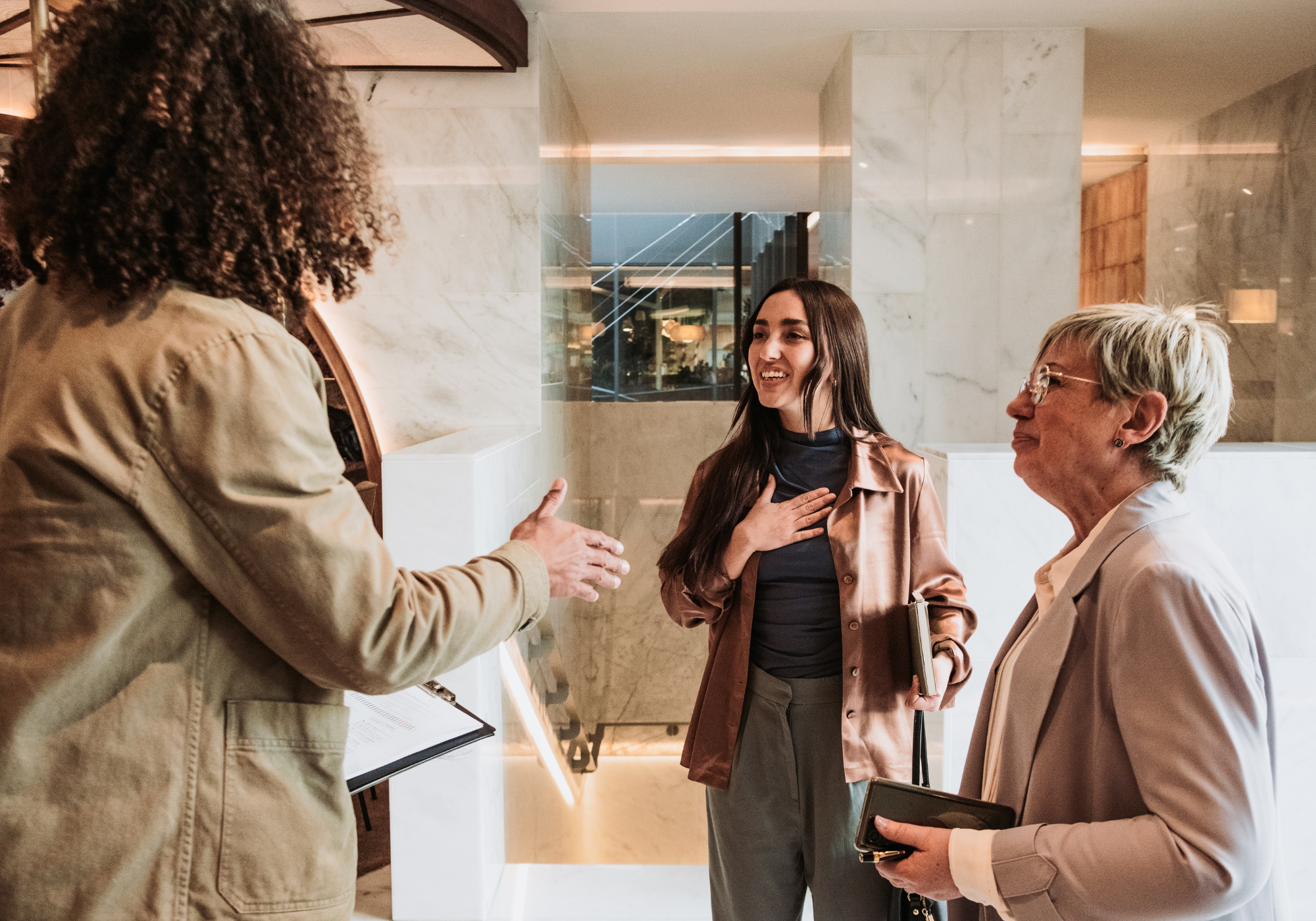 Three women engaged in conversation indoors, one woman with curly hair gesturing, a second woman with long dark hair placing her hand on her chest, and a third woman with short blonde hair holding a notebook.