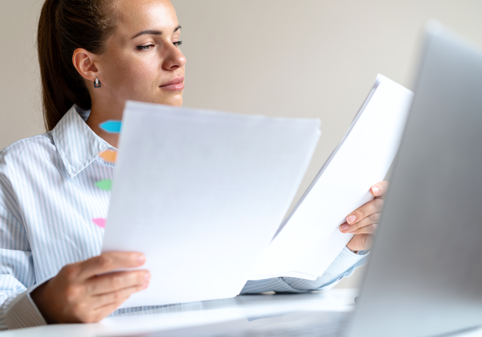 A woman with brown hair pulled back, wearing a striped shirt, sitting at a desk, reading papers with colorful tabs while working on a laptop.