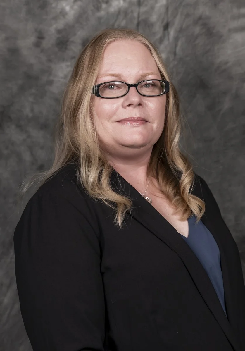 A middle-aged woman with blonde hair, wearing glasses, a black blazer, and a dark blouse, smiling softly at the camera against a gray textured background.