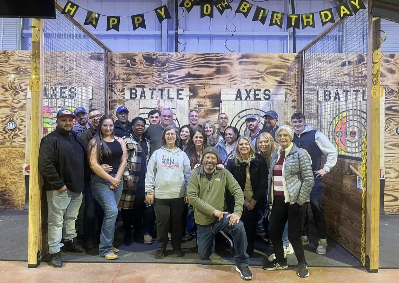 A group of people posing in front of a wooden axe-throwing target area with a 'Happy 60th Birthday' banner overhead.