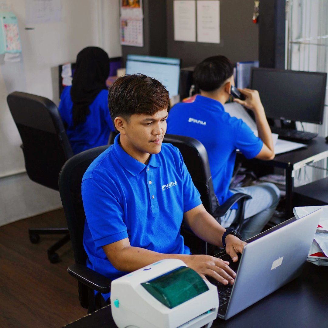 Office setting with three people working on computers, wearing blue uniforms. One person is typing on a laptop, another is speaking on the phone, and a third is facing a desktop computer. A small printer is also visible.