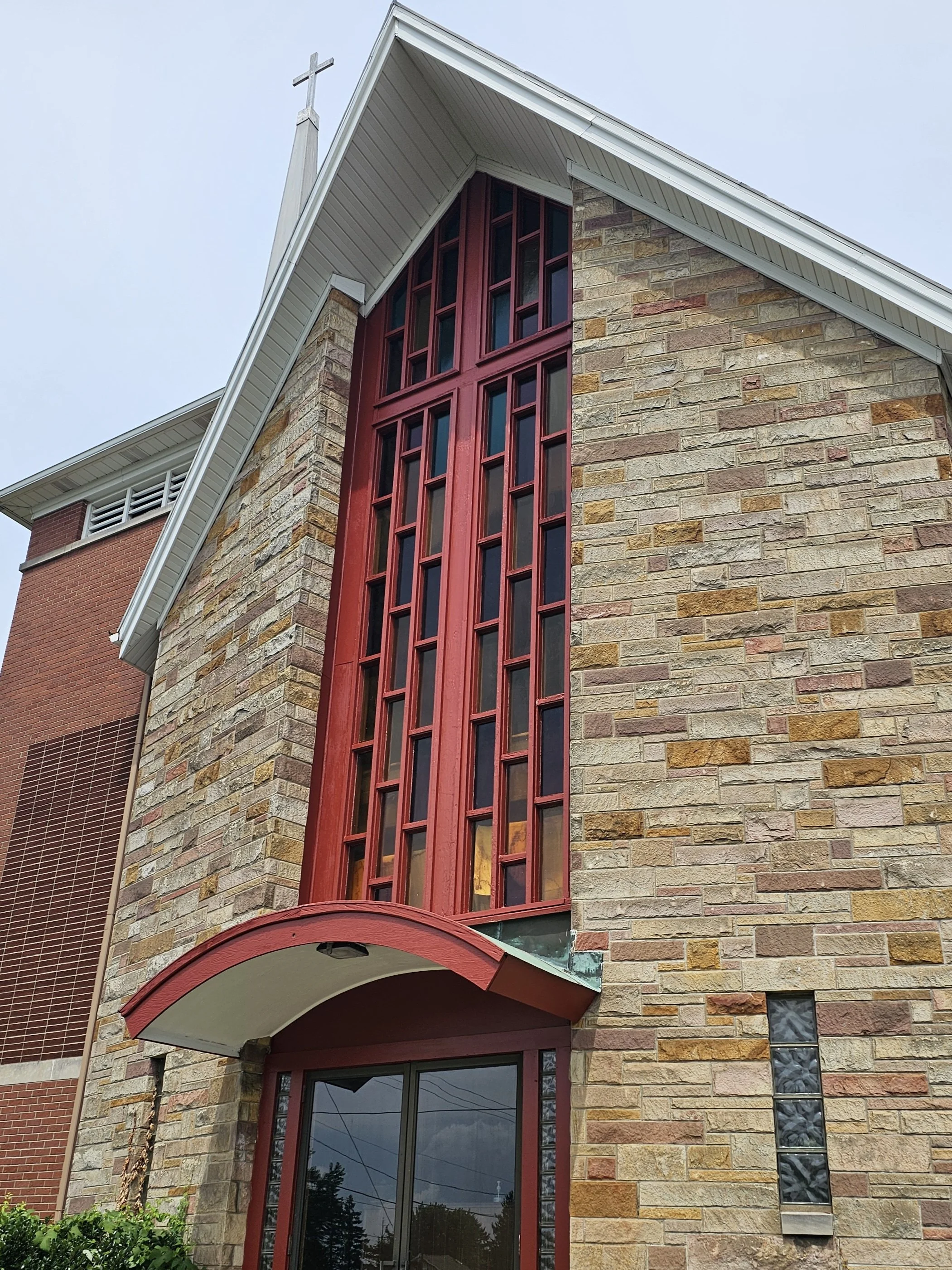 Close-up of a church building's facade with a large red stained glass window, a brick exterior with varying shades, and a small cross on top of the roof.