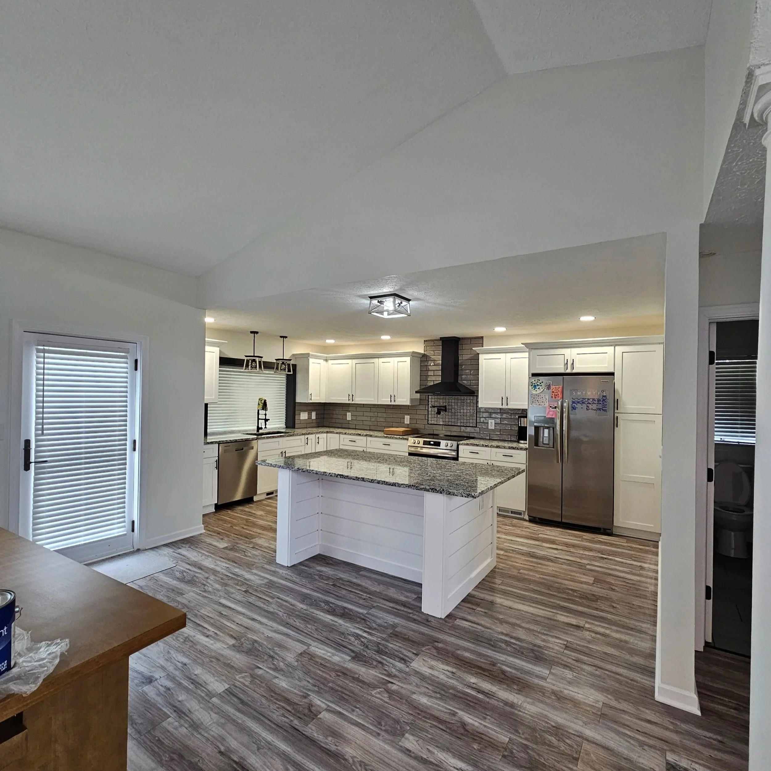 Modern kitchen with white cabinets, granite island, stainless steel fridge, black range hood, brick backsplash, and wooden flooring.