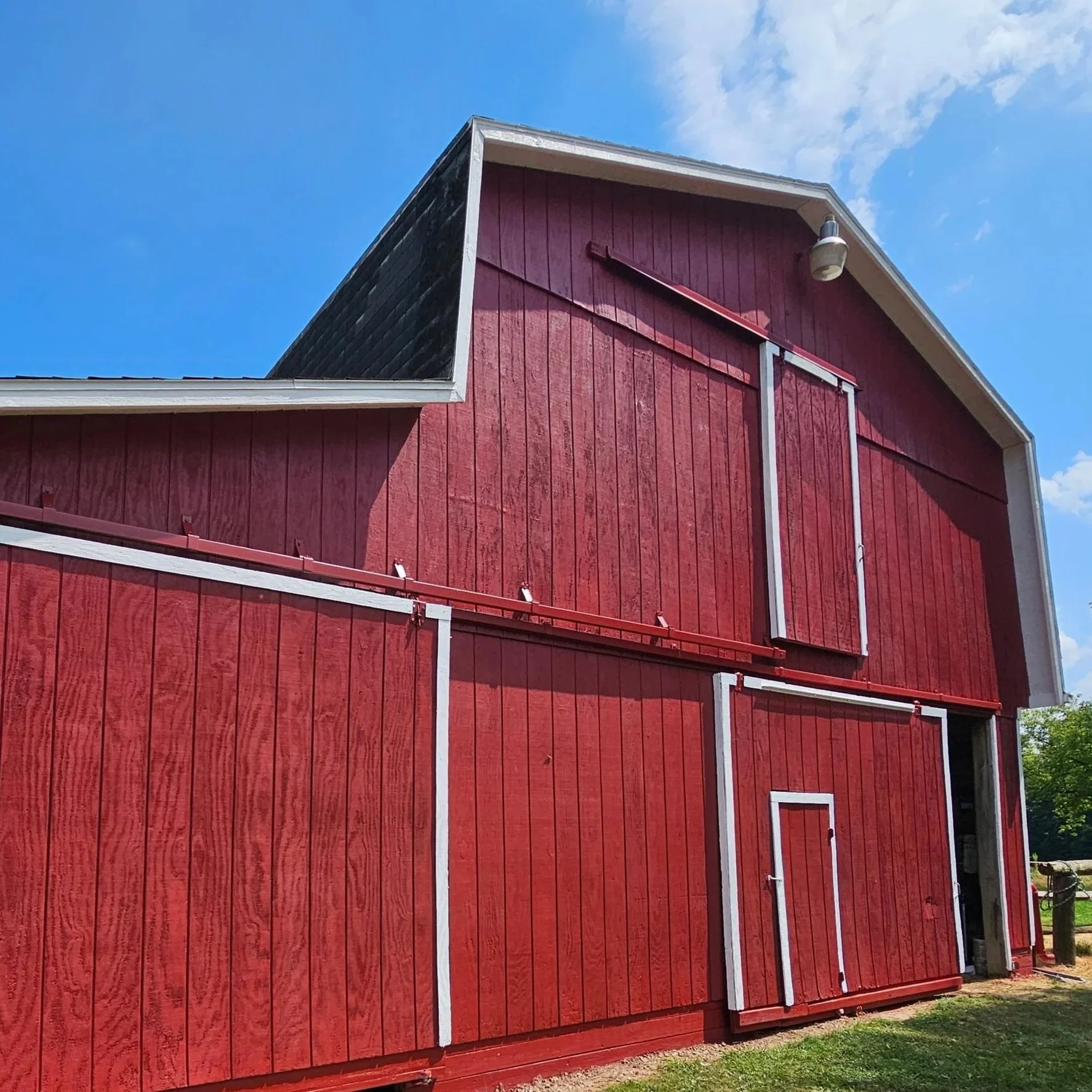 Red barn with white trim and sliding doors, under partly cloudy blue sky, surrounded by trees and grass.