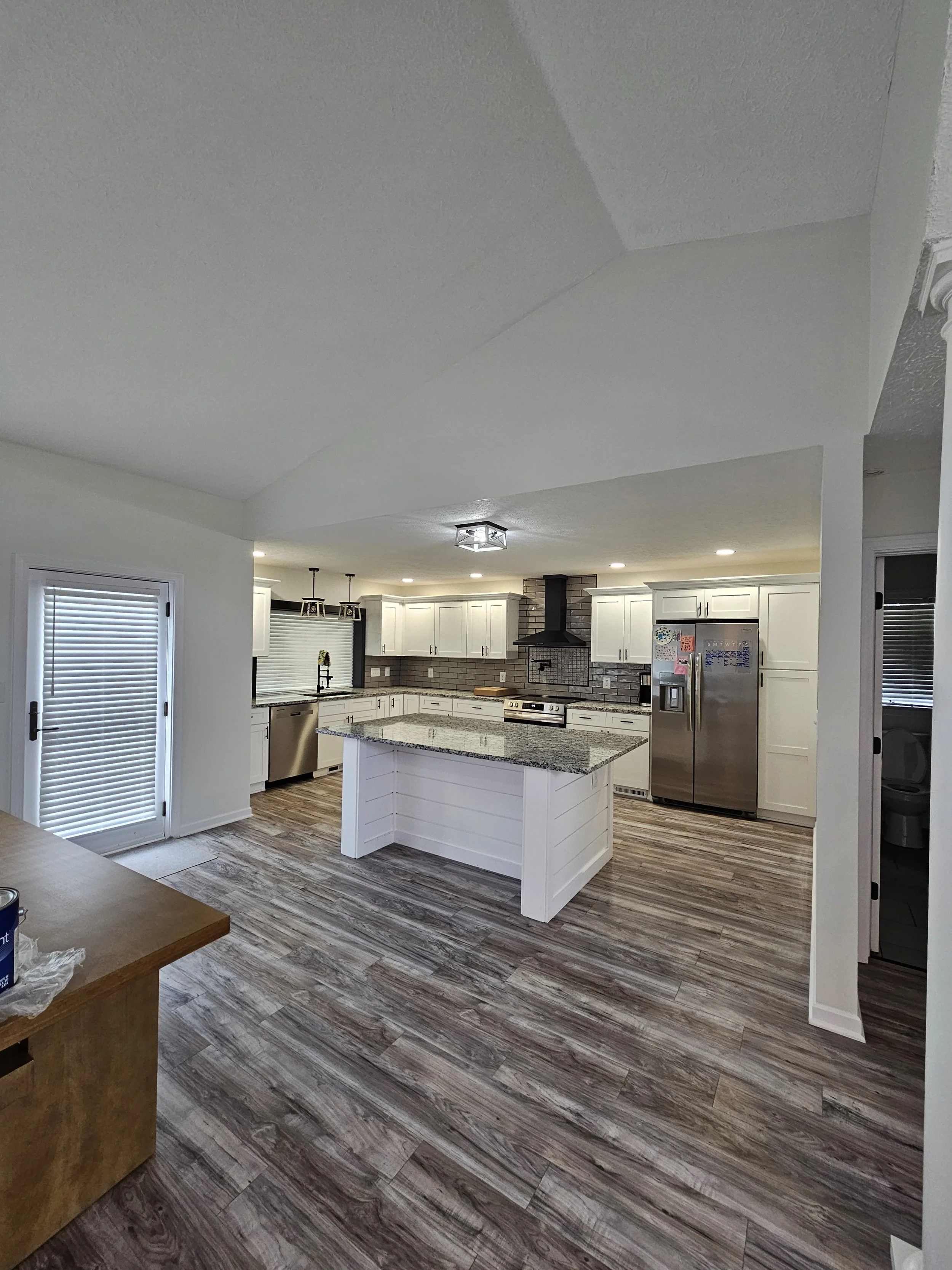 Modern kitchen with white cabinets, granite island, stainless steel fridge, black range hood, brick backsplash, and wooden flooring.