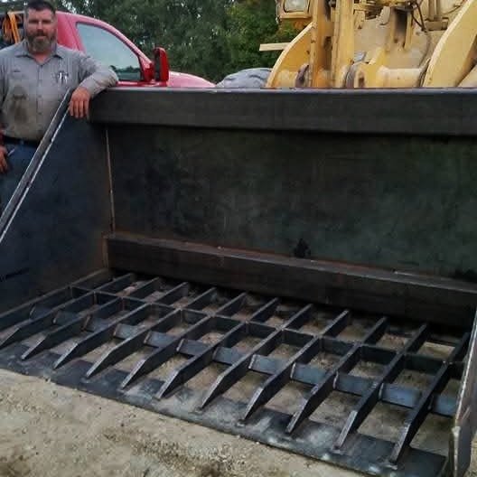 Man standing next to a large metal industrial attachment with a grated surface, likely part of construction equipment, on a dirt area with vehicles in the background.