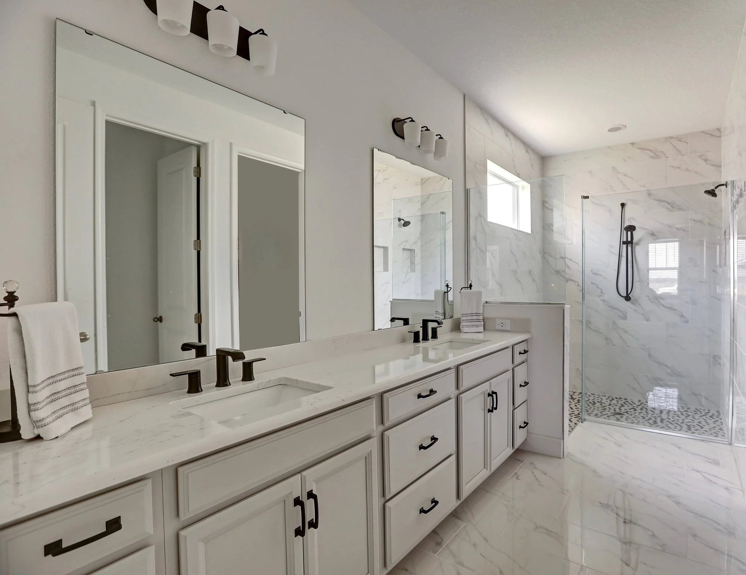 Modern bathroom with white cabinetry, dual sinks, large mirrors, marble countertops, wall-mounted black faucets, towel rack, and walk-in shower with marble walls, black fixtures, and pebble tile floor