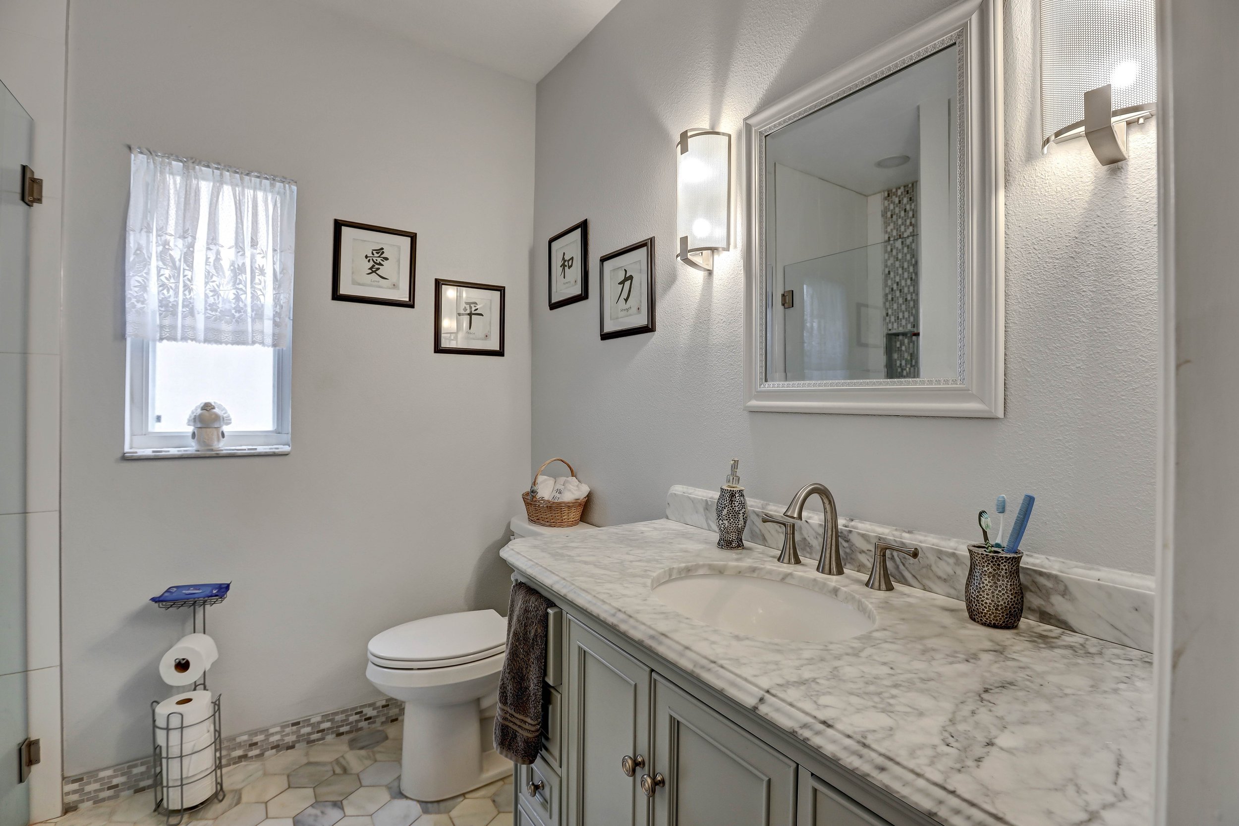 A small bathroom with a window, framed artwork on the wall, a mirror above a marble countertop with a sink, a toothbrush holder, and a soap dispenser. A towel hangs from the cabinet, and a toilet is next to the vanity. The floor has hexagonal tiles.