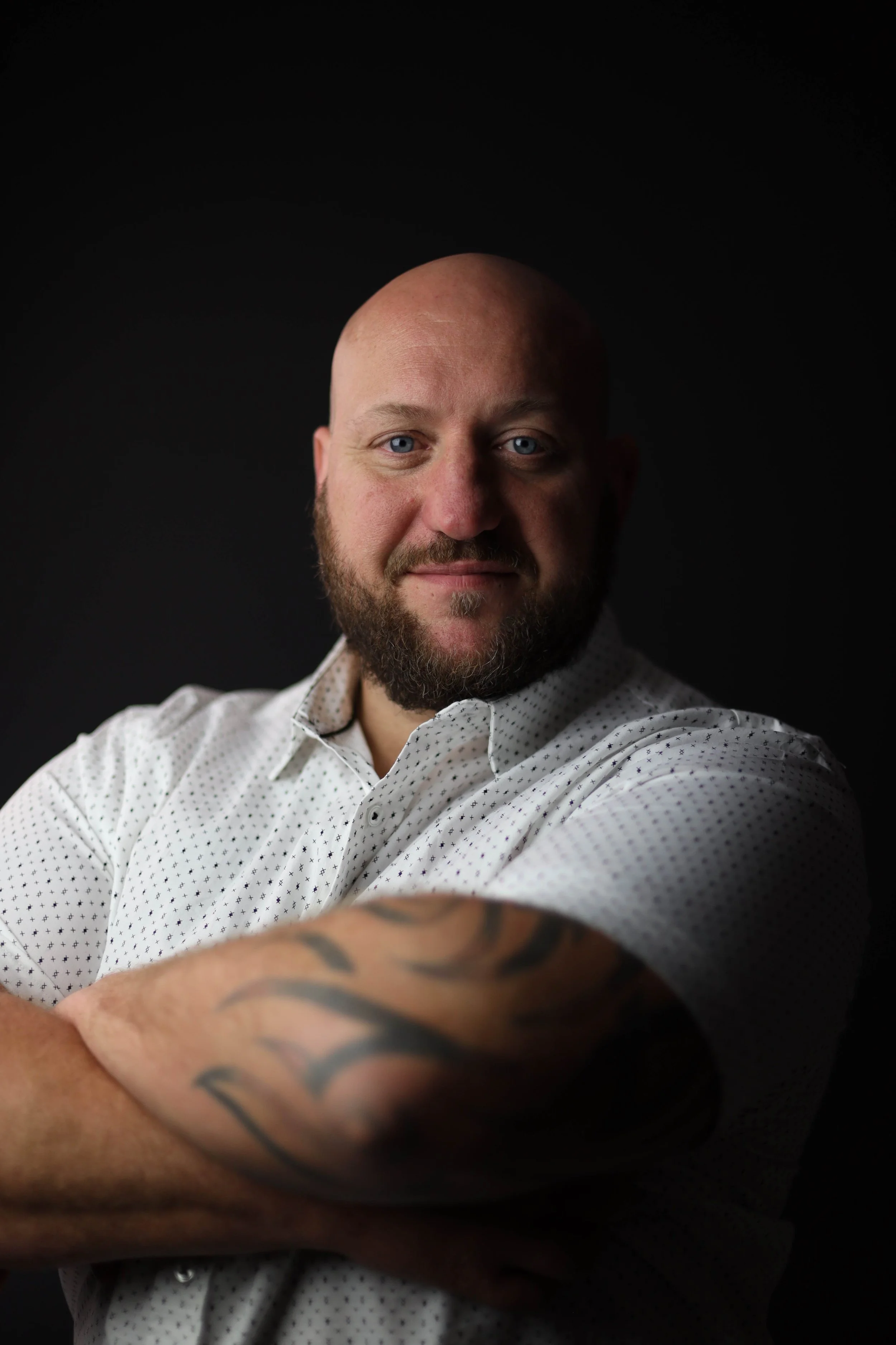 A man with a beard and bald head, wearing a white dotted shirt, smiling, with arms crossed, standing against a dark background.