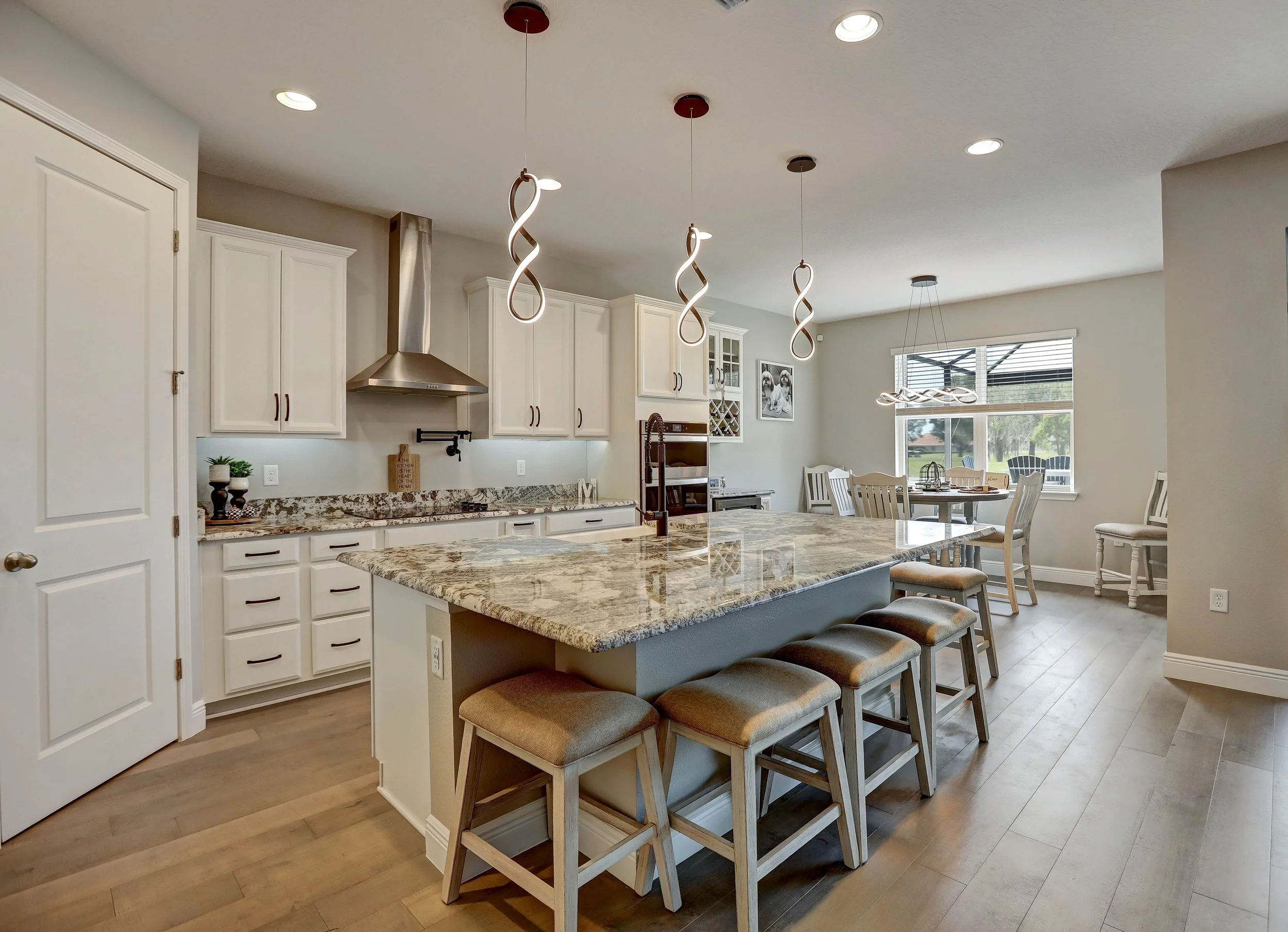 A modern kitchen with white cabinets, a granite countertop, and a large island with barstools. There are three hanging light fixtures above the island, a stove with a stainless steel range hood, and a dining area with a table and chairs near a window