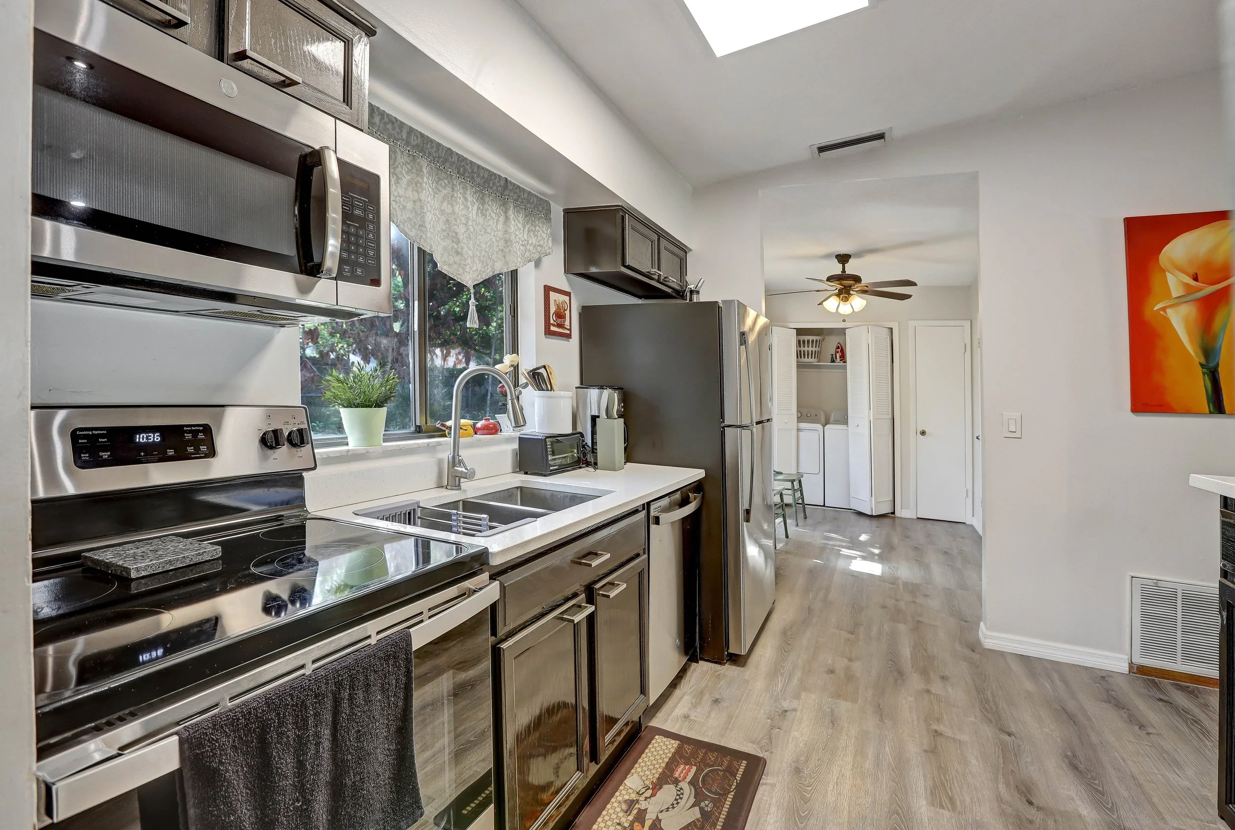 Kitchen with stainless steel microwave, stove, refrigerator, and white countertops. There is a window above the sink, dark cabinets, and a ceiling fan in the adjacent laundry area.